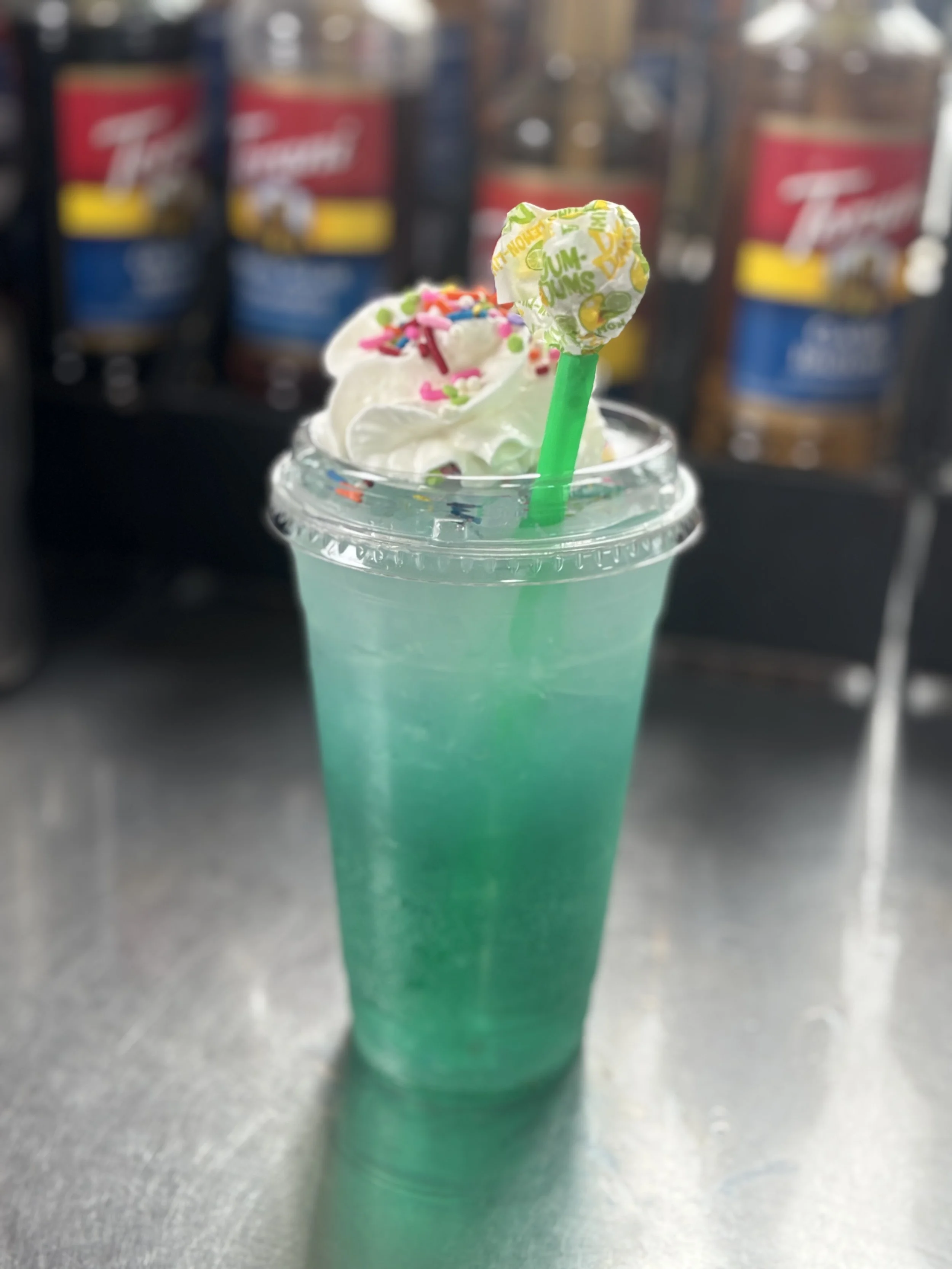 A blue slushy drink topped with whipped cream, colorful sprinkles, and multicolored edible glitter, garnished with a green straw and a lemon-lime flavored lollipop, on a metallic surface with a vending machine in the background.