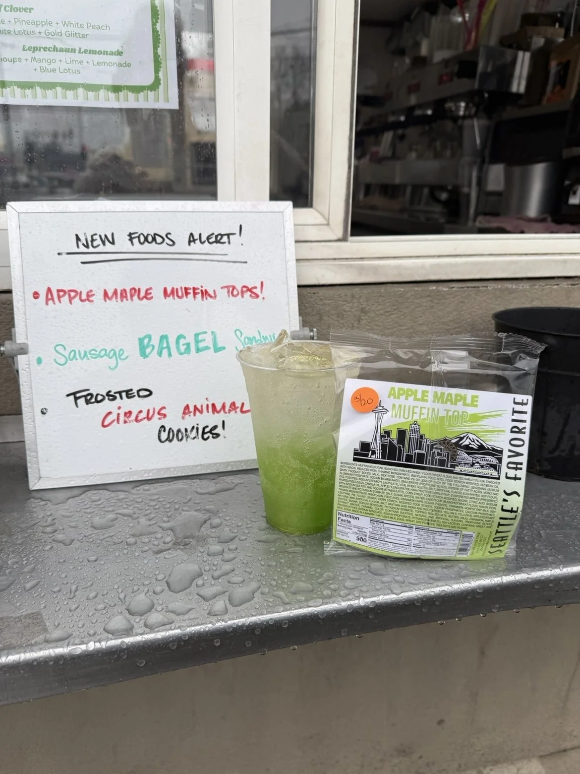 A signboard with new food alerts, including apple maple muffin tops, sausage bagel, and frosted circus animal cookies. A green drink with ice in a plastic cup, and a package of apple maple muffin tops labeled 'favorite' from Seattle, with raindrops o