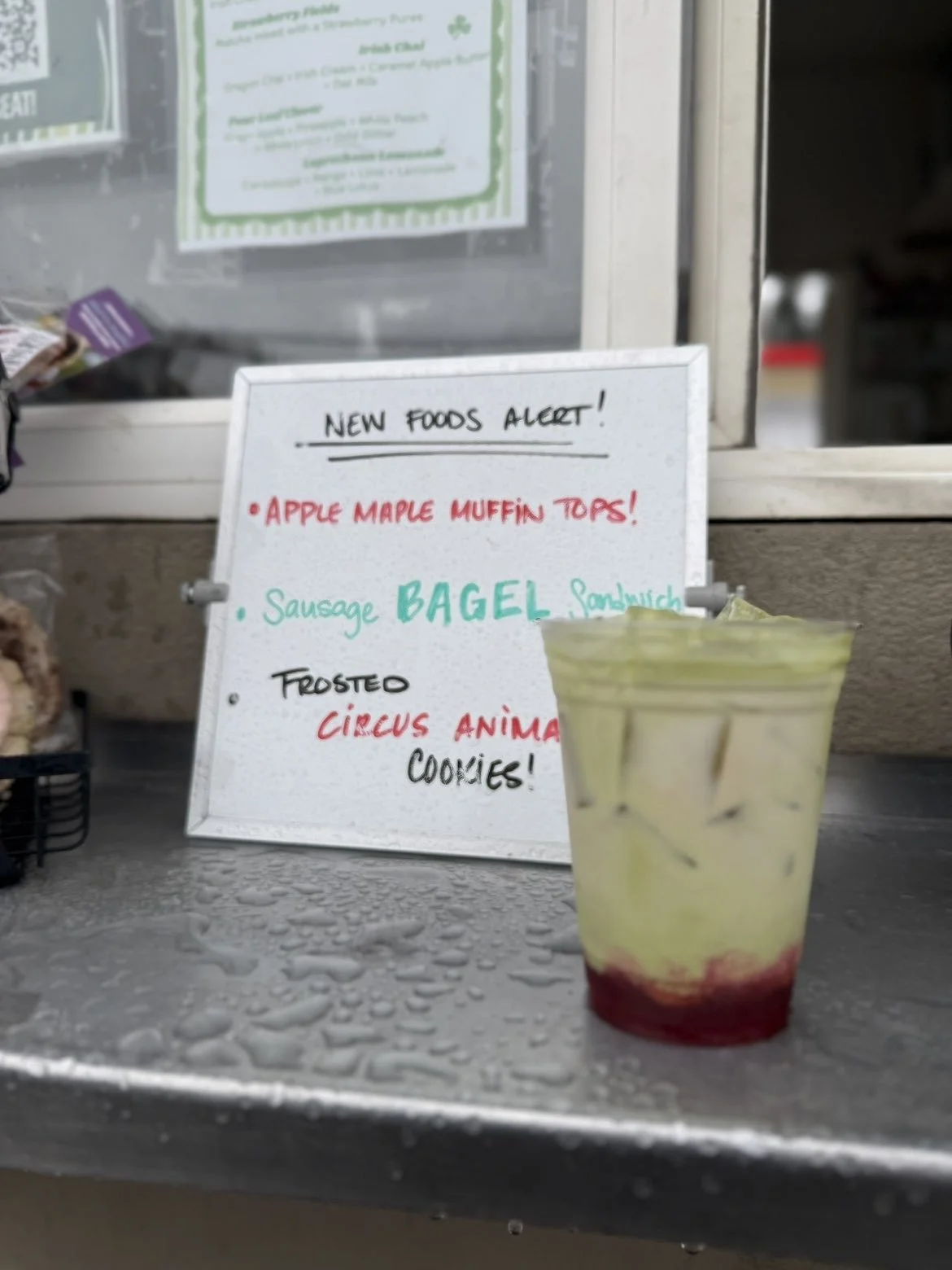 Signboard displaying new food items including apple maple muffin tops, sausage bagel sandwich, and frosted circus animal cookies, with a drink in front.