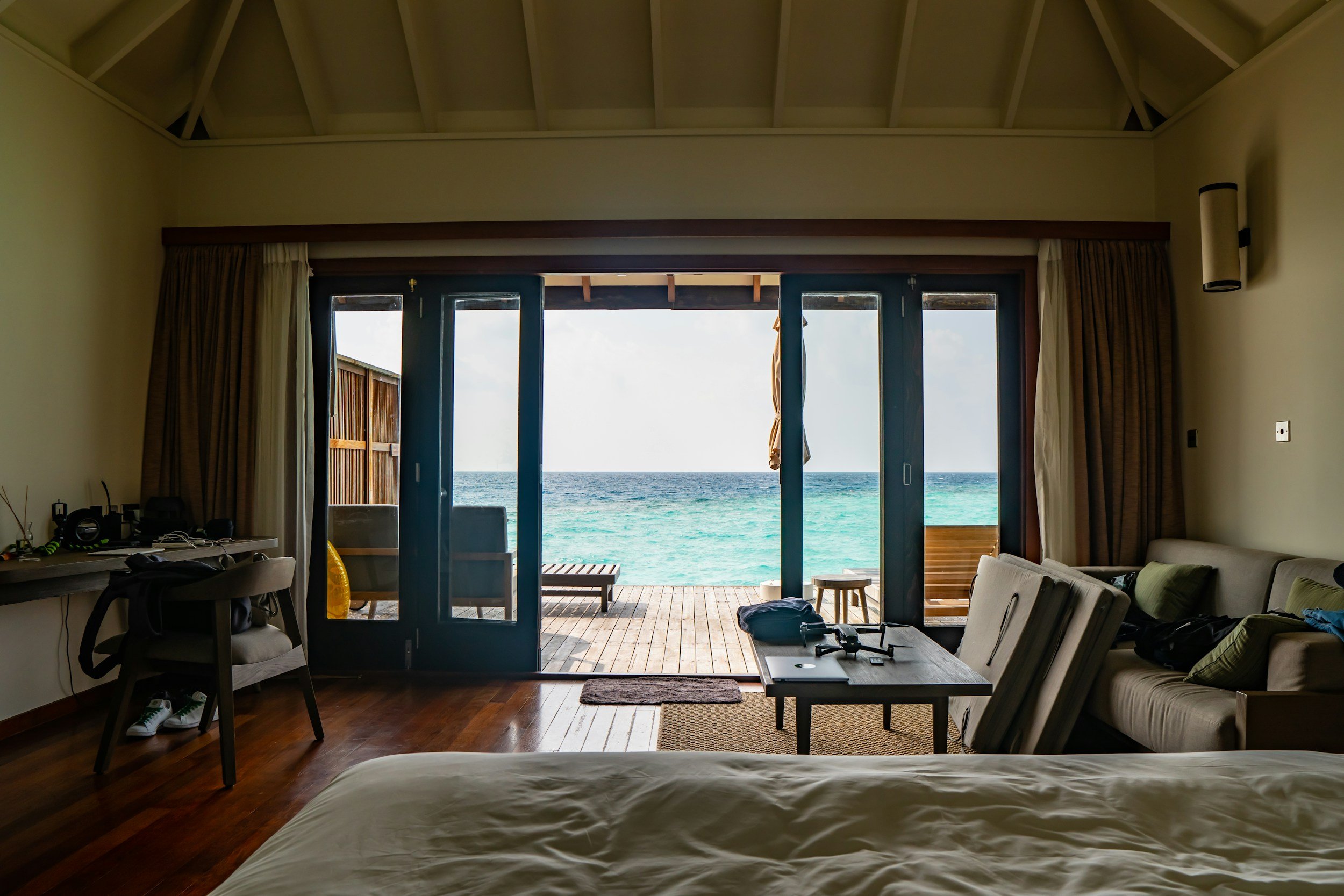 Interior of a bedroom with large glass doors opening to a balcony facing the ocean, with chairs and a small table outside, natural light streaming in.