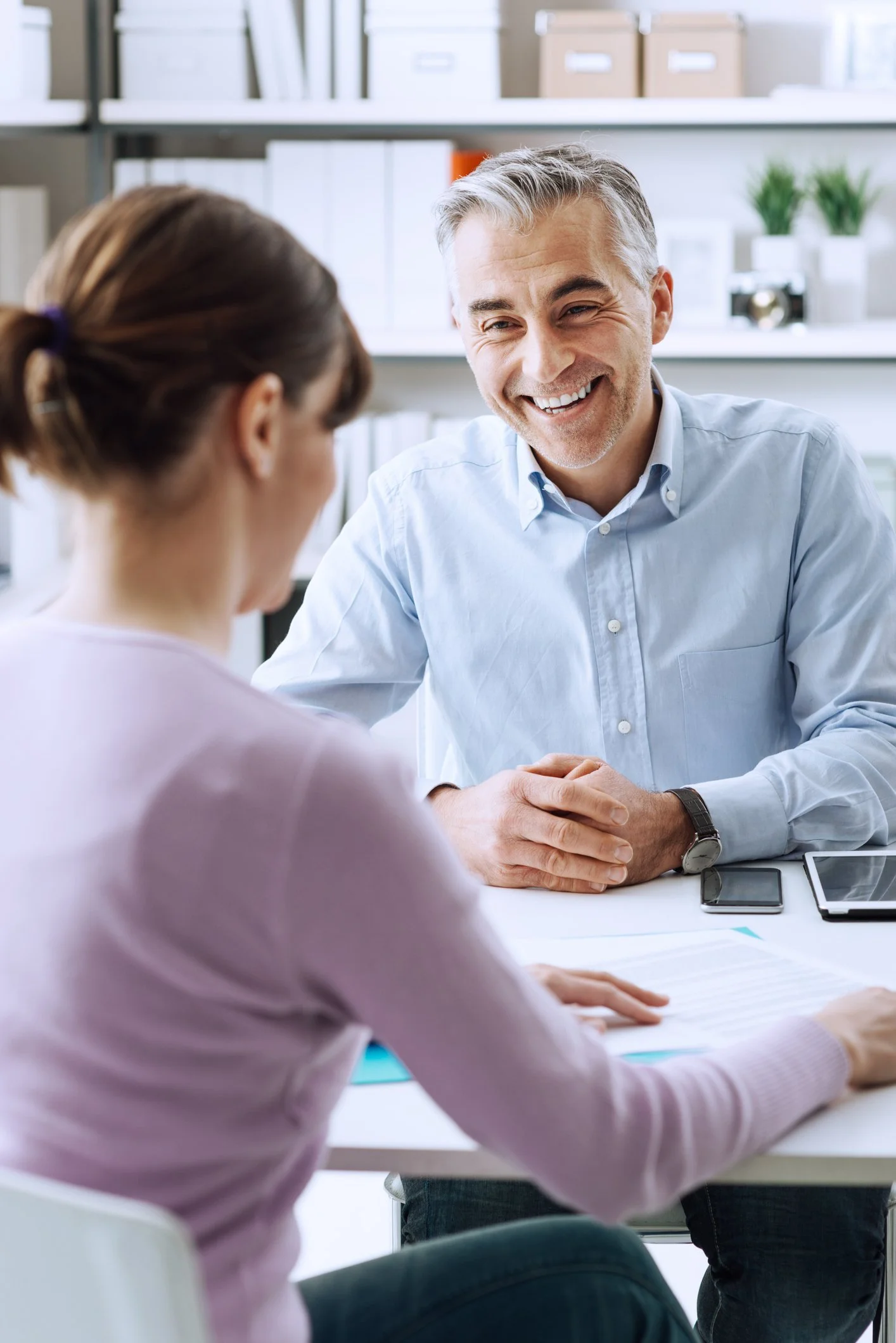 A man in a blue shirt, a San Jose Medicare insurance agent, smiles while talking to a woman across a desk in a bright office.