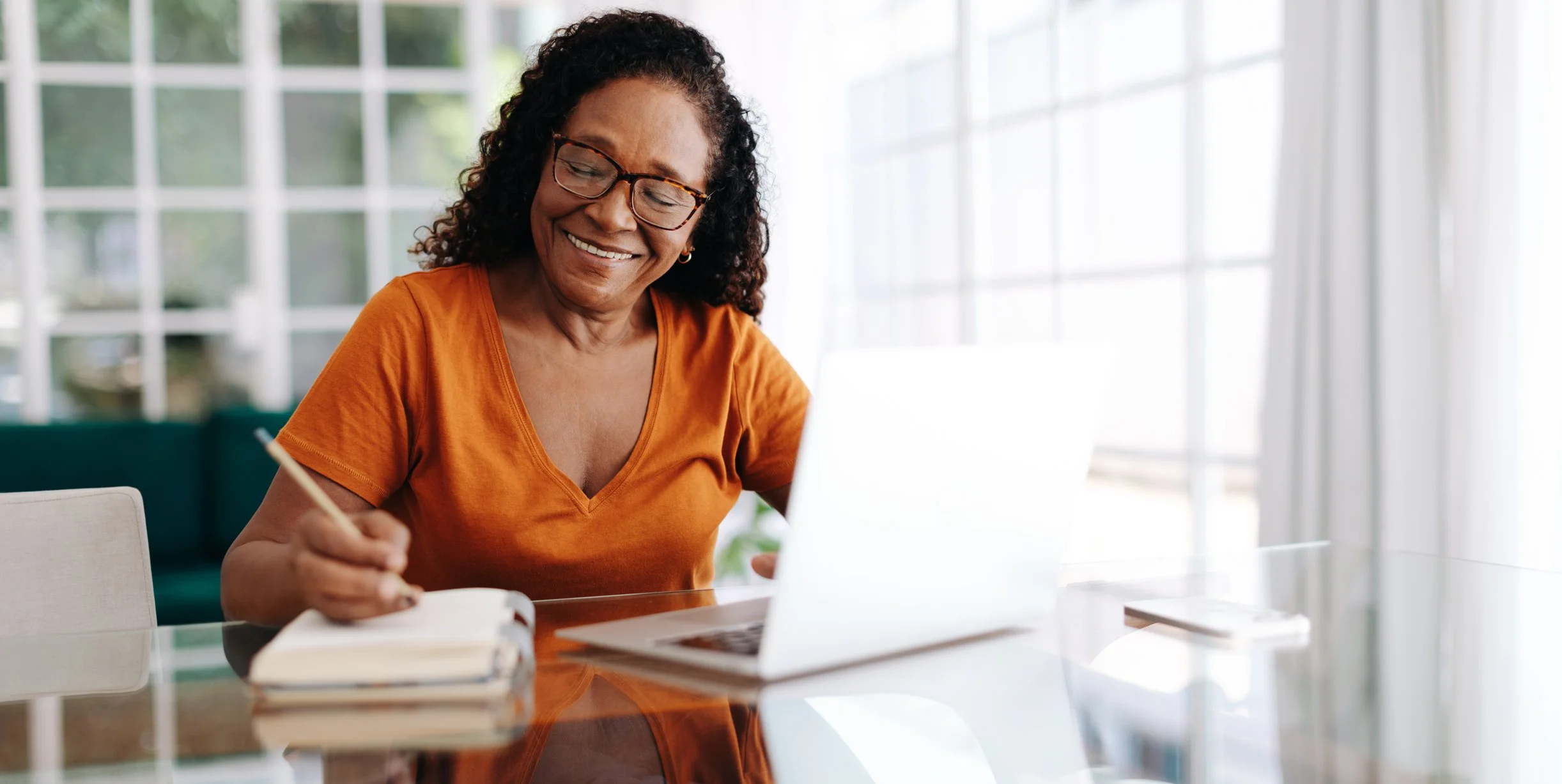 Smiling woman in orange shirt researches Medicare Part D as she writes in a notebook and uses a laptop at a bright glass table.