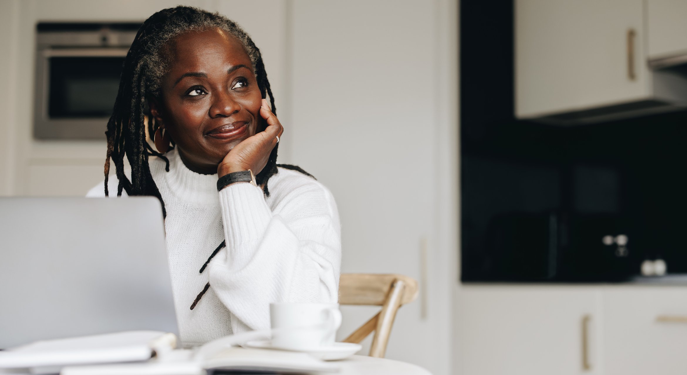 Smiling woman with gray braids sits at a table, pondering Medicare Advantage Insurance as she sips coffee beside her laptop.