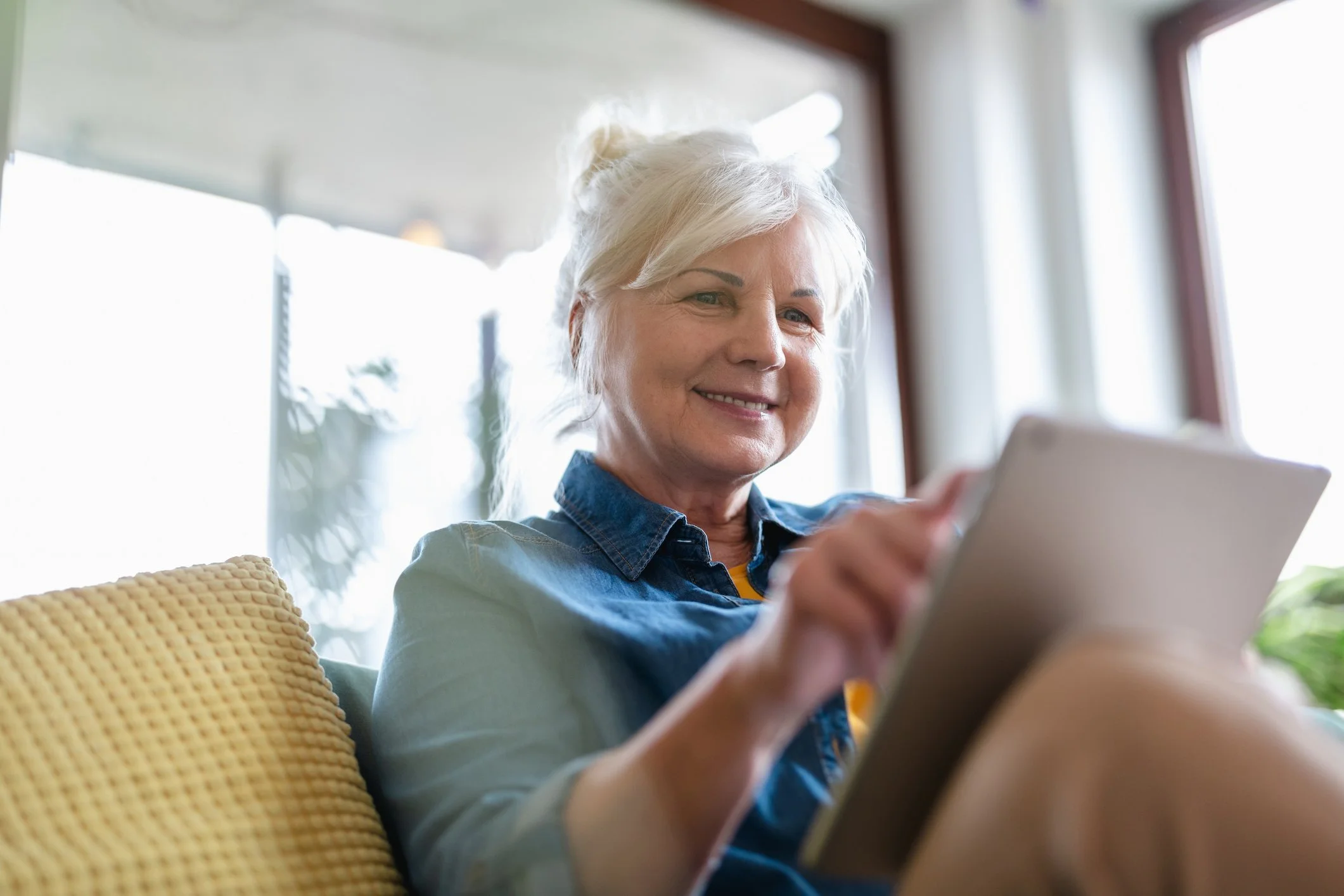 Smiling older woman sits on a couch using a tablet, researching Medicare Advantage Insurance as sunlight streams in behind her.