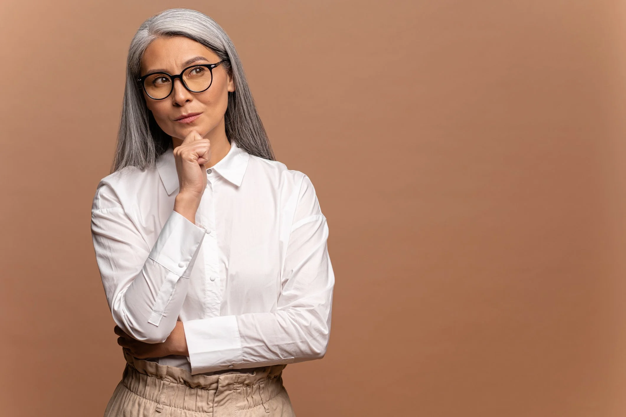 Woman with long gray hair and glasses in white shirt, looking thoughtful, perhaps considering a Medicare Supplement Insurance Plan.