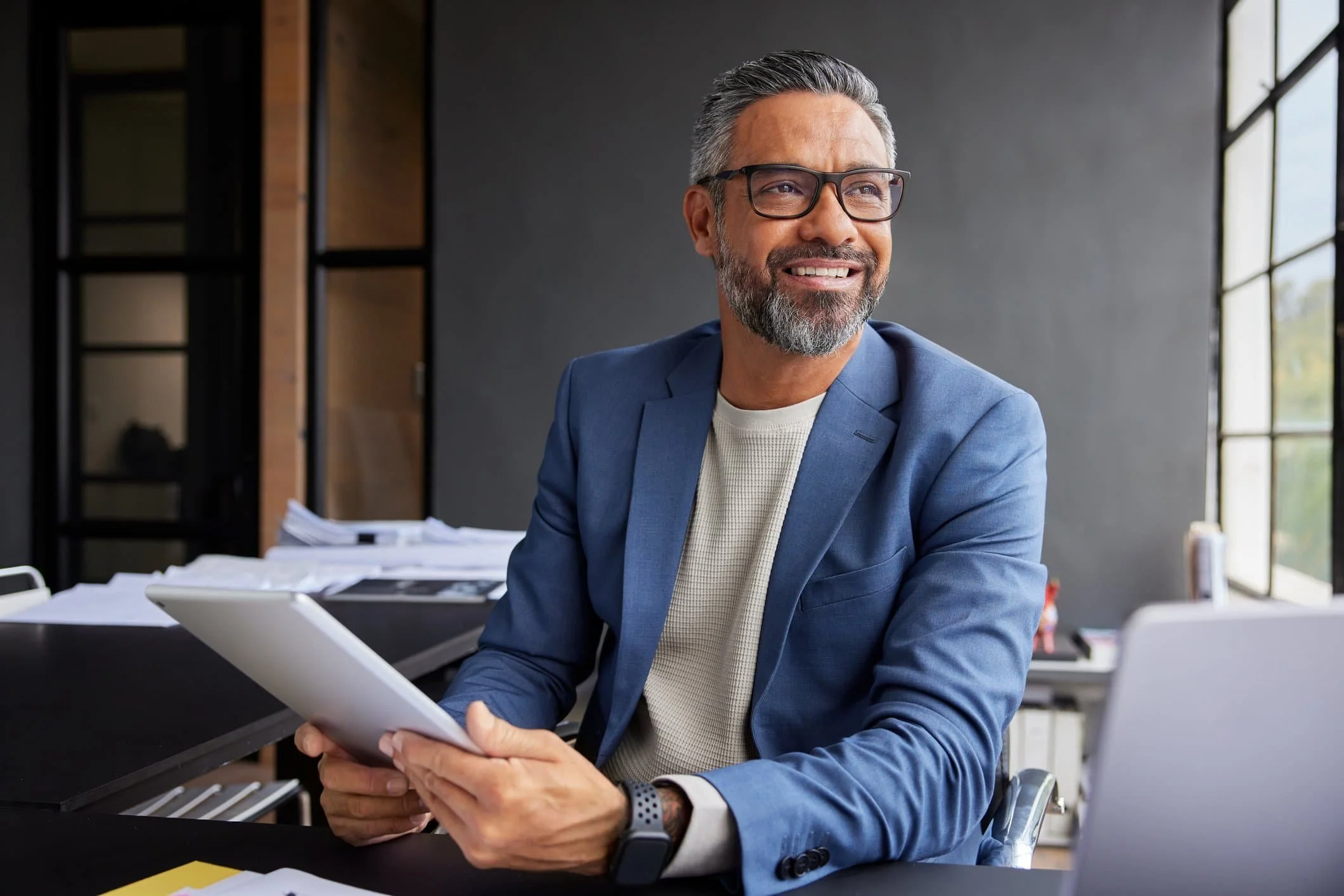 Smiling man in a blue blazer sits at a desk offering Insurance Guidance, holding a tablet with papers and laptop in a modern office.