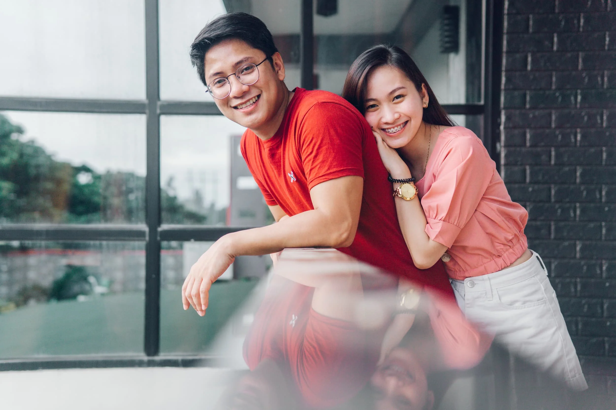 Smiling man in red shirt and woman in pink shirt lean together by a window, discussing life insurance; their reflections shine below.