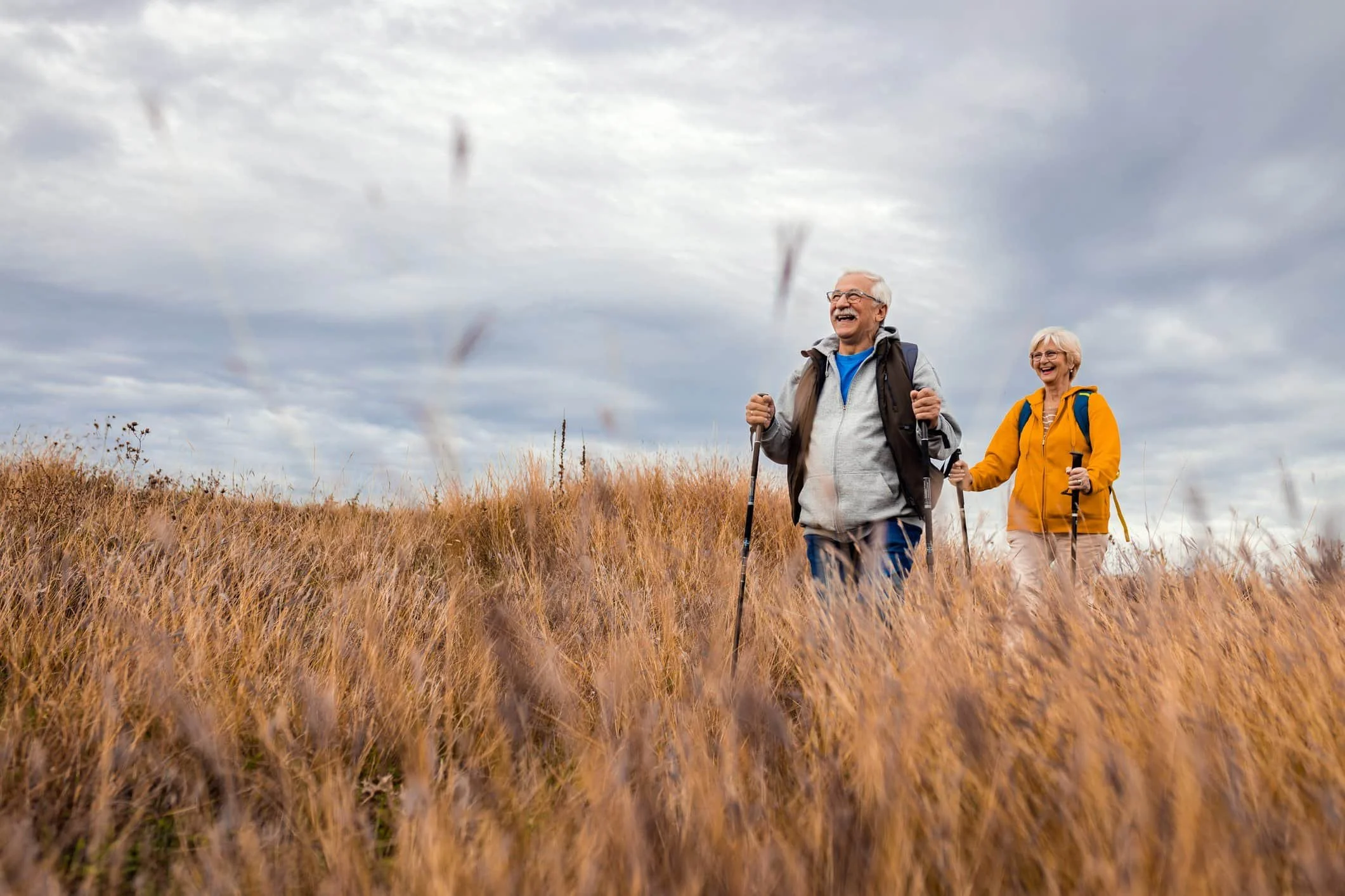 Two older adults, turning 65, hike through tall grass under a cloudy sky, smiling and using trekking poles.