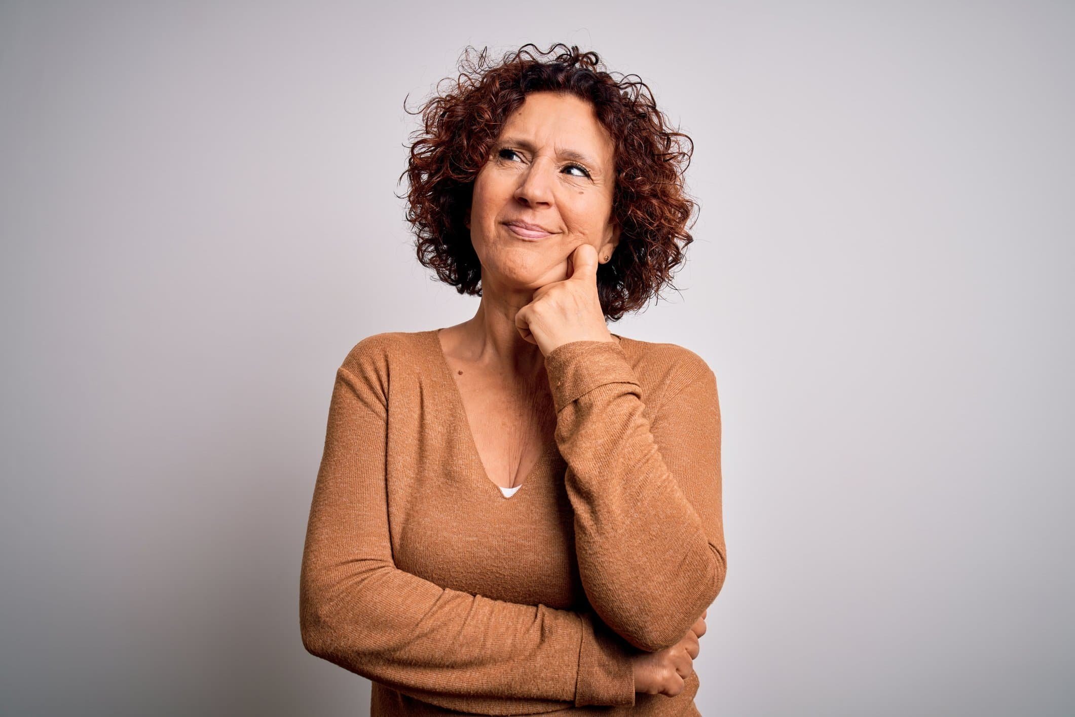 Curly-haired woman in a brown sweater smiles thoughtfully, hand on chin, reflecting on turning 65 and signing up for Medicare soon.