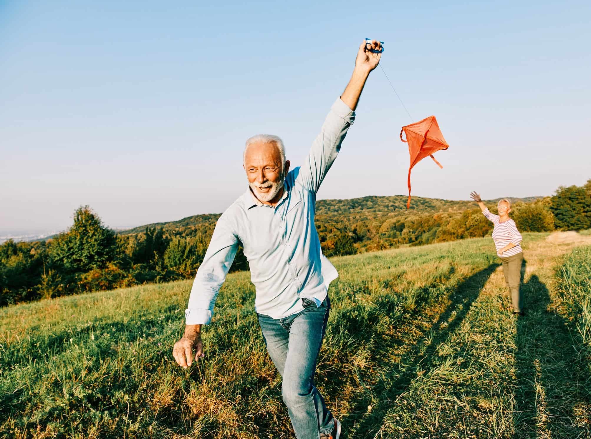 Smiling older man runs with a red kite in a grassy field, enjoying life and Medicare Advantage Insurance on a sunny day.