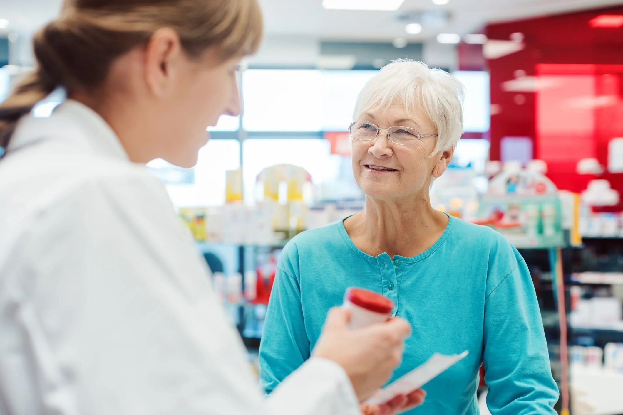 An older woman smiles while talking to a pharmacist about her Medicare Part D prescription in a brightly lit pharmacy.