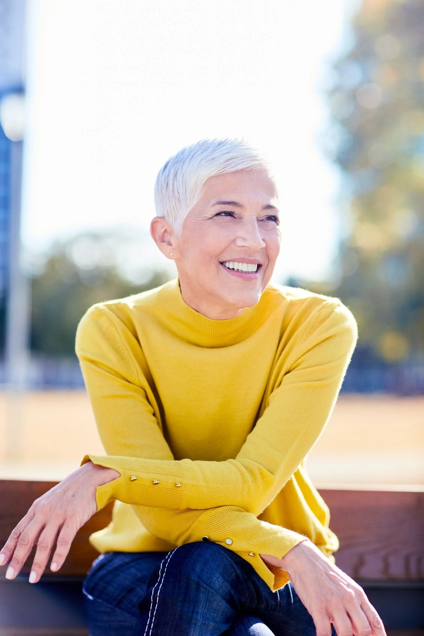 Smiling woman with short gray hair in a yellow sweater sits outside on a bench, enjoying a sunny day near her Medicare Insurance Agency.