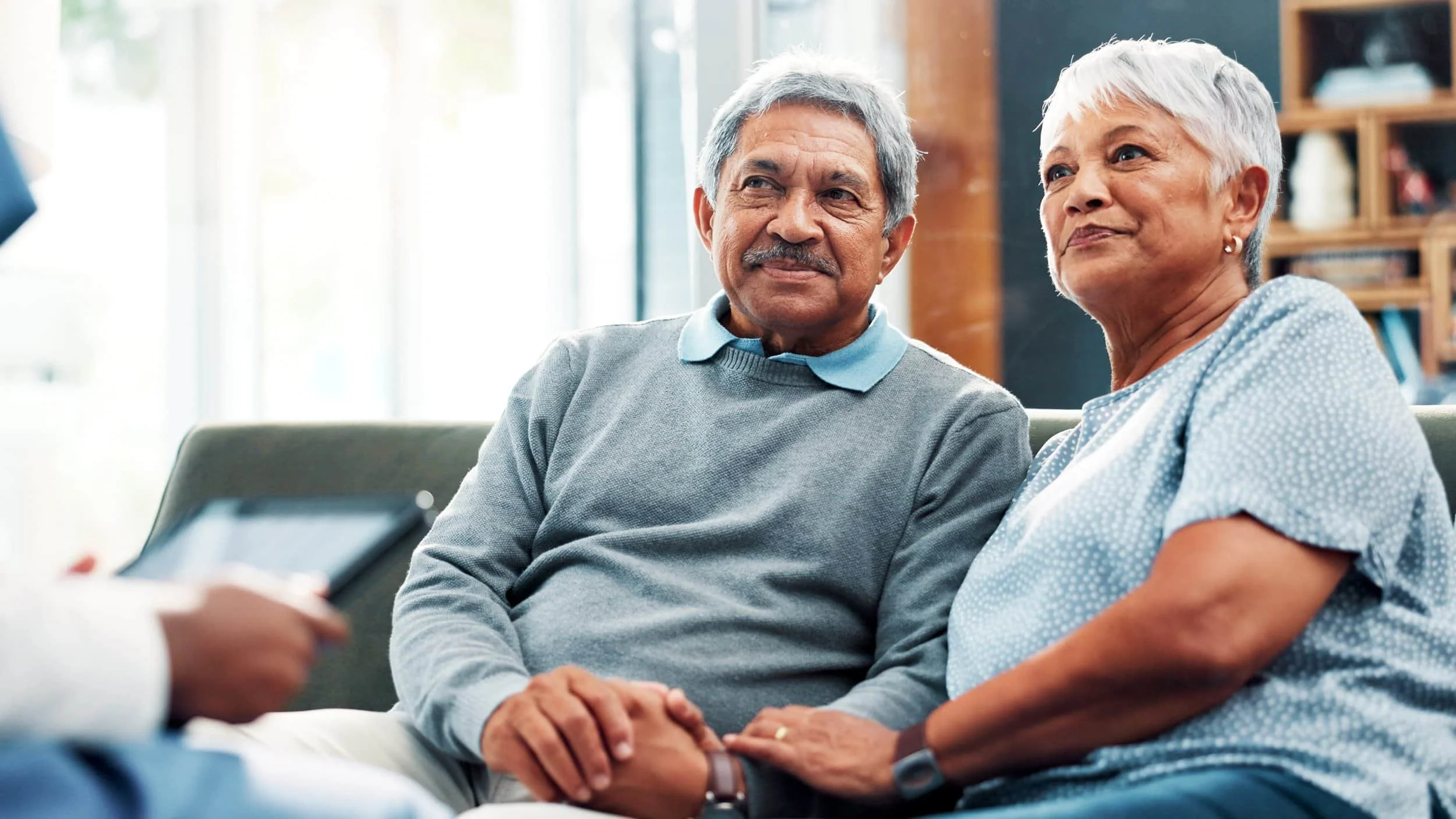 Older couple sits on a couch, smiling and holding hands, discussing options with a Medicare Insurance Agency representative off camera.