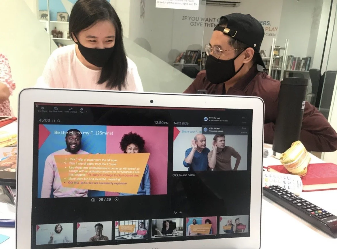 Two people, one woman and one man, wearing face masks and glasses, are having a discussion at a table with a laptop displaying a presentation.