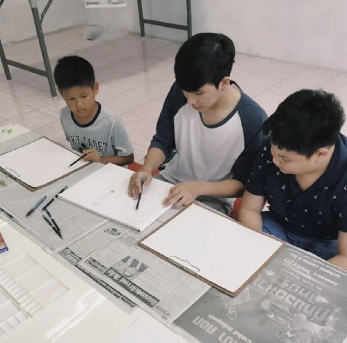 Three boys sitting at a table with blank boards, newspapers, and drawing tools, engaged in an art activity indoors.