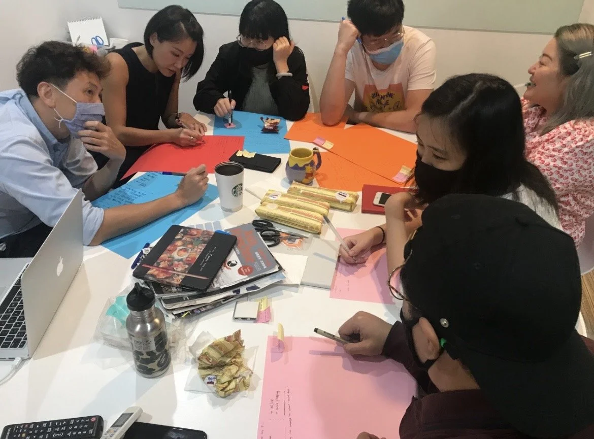 Group of people sitting around a table engaged in a discussion or activity, with various papers, notebooks, pens, masks, and drinks on the table.