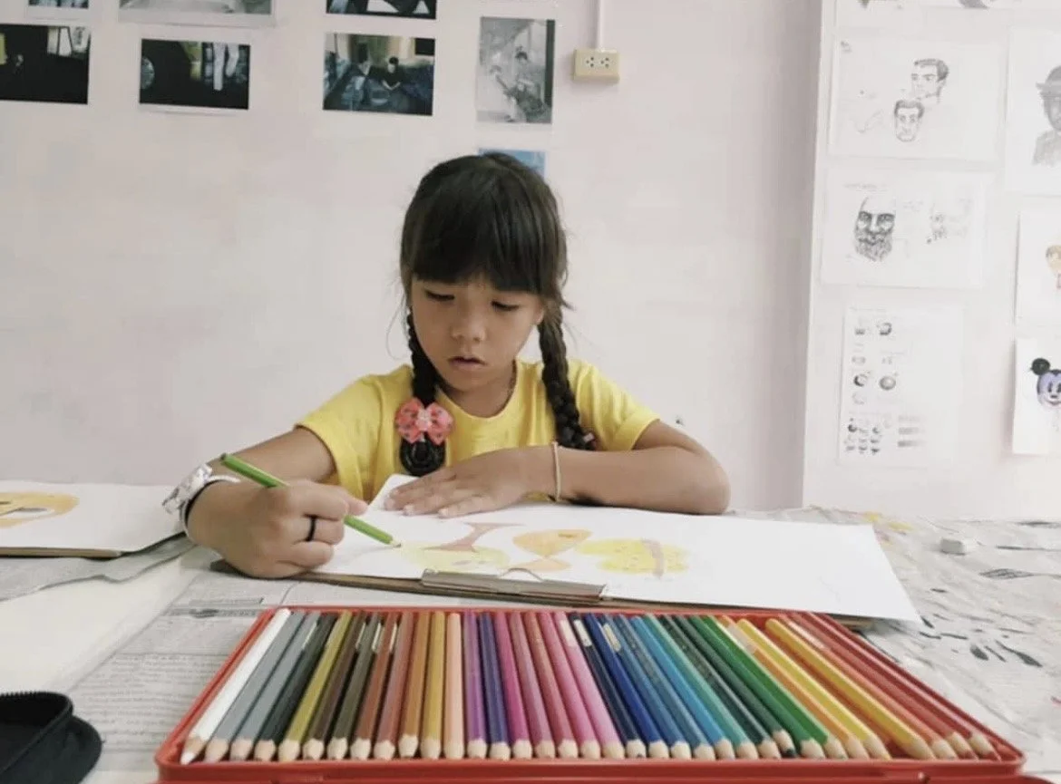 Young girl with braided hair and pink bows, wearing a yellow shirt, drawing with colored pencils at a table with a large set of colored pencils, in a room decorated with photographs and sketches.