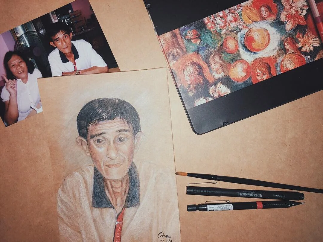 A collection of art supplies and personal photos on a wooden surface. There is a detailed colored pencil drawing of a man, a photograph of two children smiling, a tablet displaying an abstract painting, and three pens.