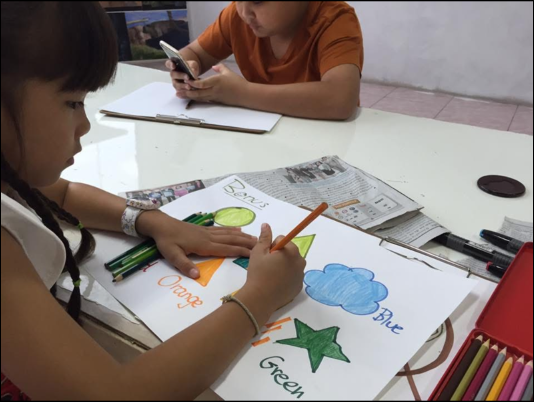 Child drawing colorful shapes and words on paper, with an adult using a smartphone and various art supplies on the table.