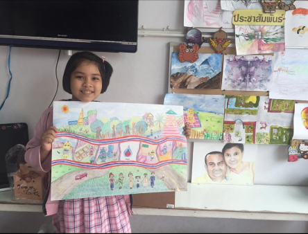 A young girl in a pink and white checkered dress holding up a colorful drawing of a landscape with houses, trees, and a rainbow. There are various artworks displayed on a wall behind her, including paintings and drawings of different scenes and portr