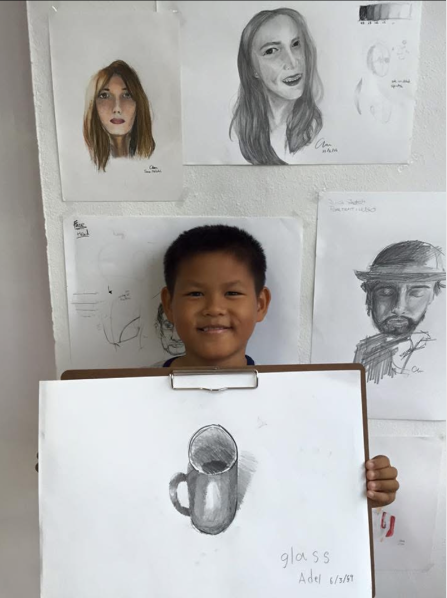 Young boy smiling while holding a drawing of a glass mug on a whiteboard, with various portrait sketches and drawings hanging on the wall behind him.