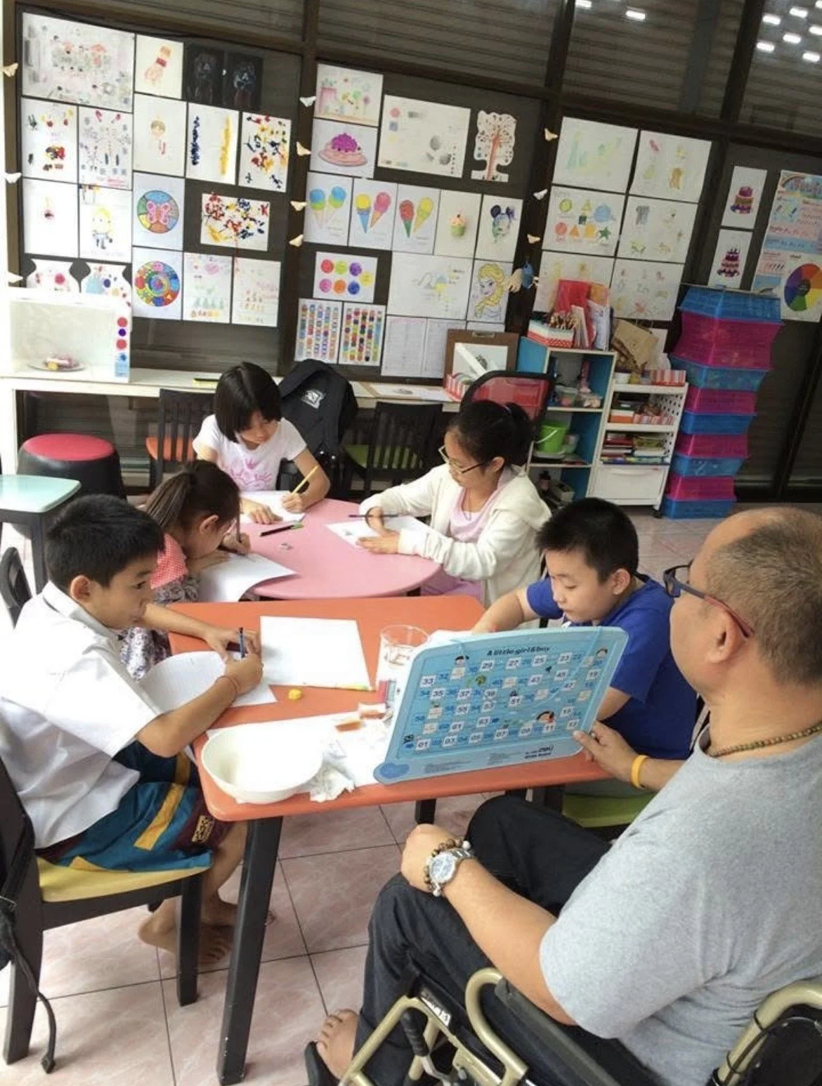 A group of children and two adults sitting around tables engaging in arts and crafts activities in a classroom decorated with colorful drawings and artwork on the walls.