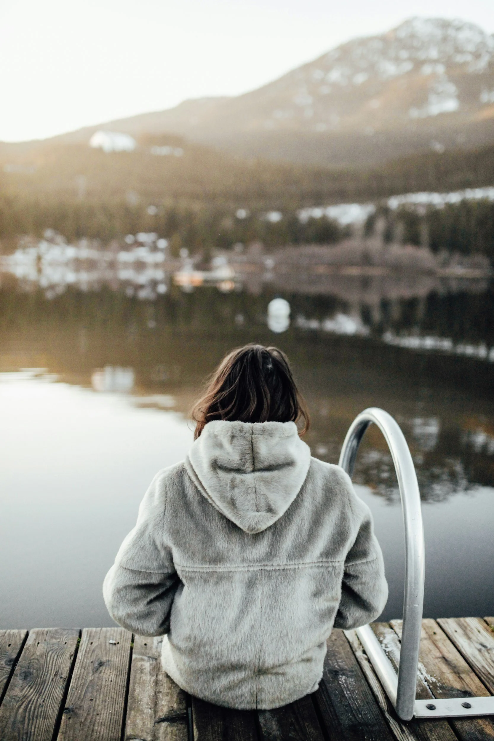 Person with long dark hair wearing a light-colored hoodie sitting on a wooden dock by a calm body of water, facing away, with mountains in the background during sunset or sunrise.