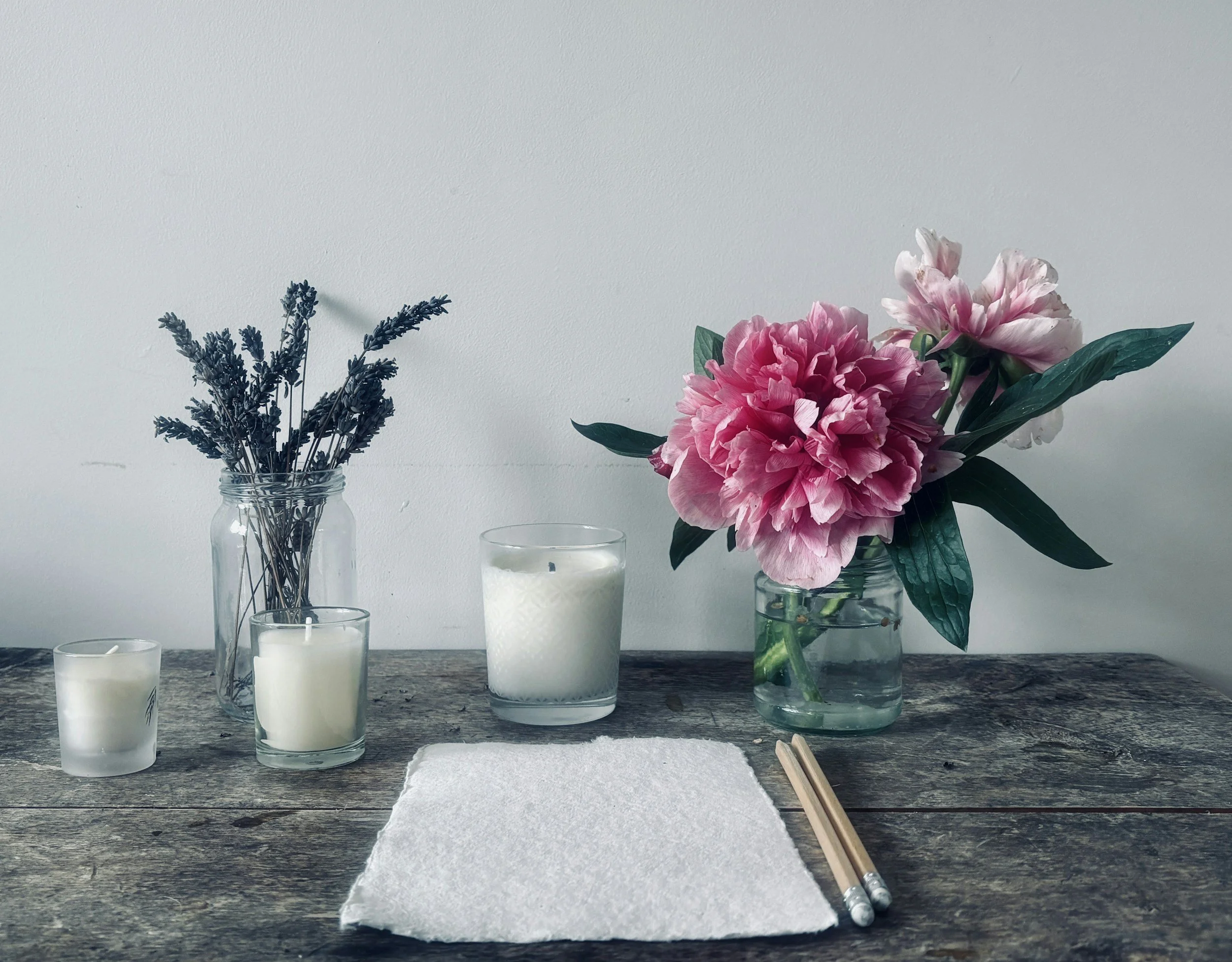 A wooden table with three white candles in glass holders, a small glass jar with lavender stems, a larger jar with pink and white peony flowers, and a pair of chopsticks on a piece of white paper.