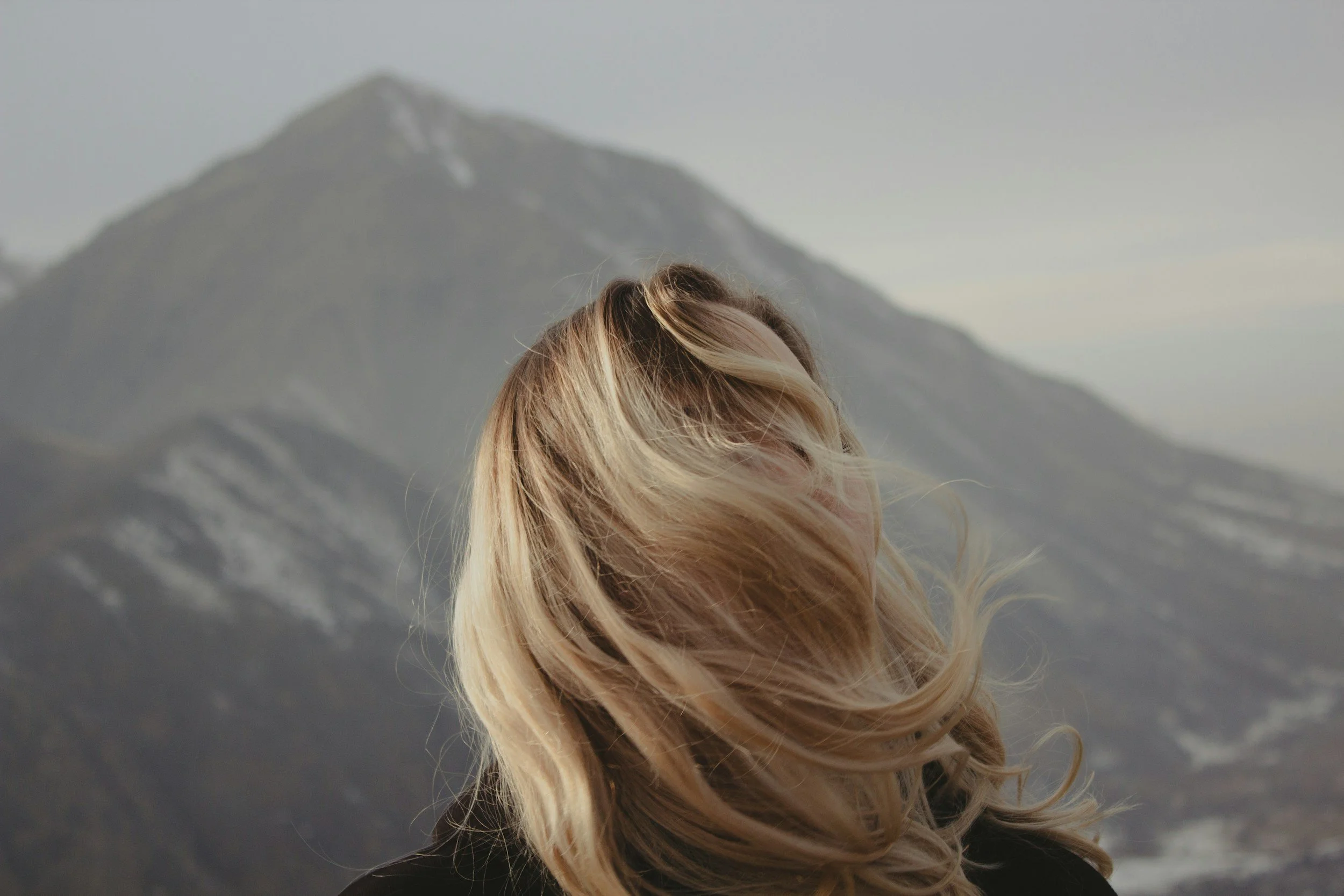 Close-up of a person with long blonde hair outdoors, with a snowy mountain in the background.