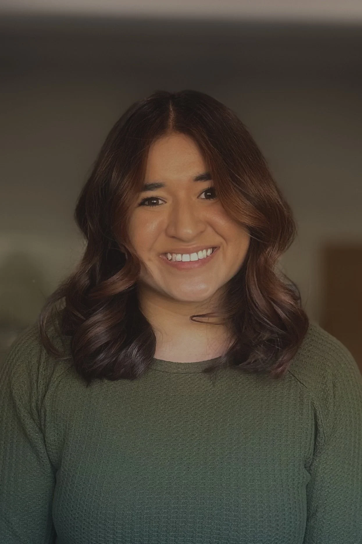 A woman with wavy, shoulder-length brown hair, smiling, wearing an olive green sweater, standing indoors.