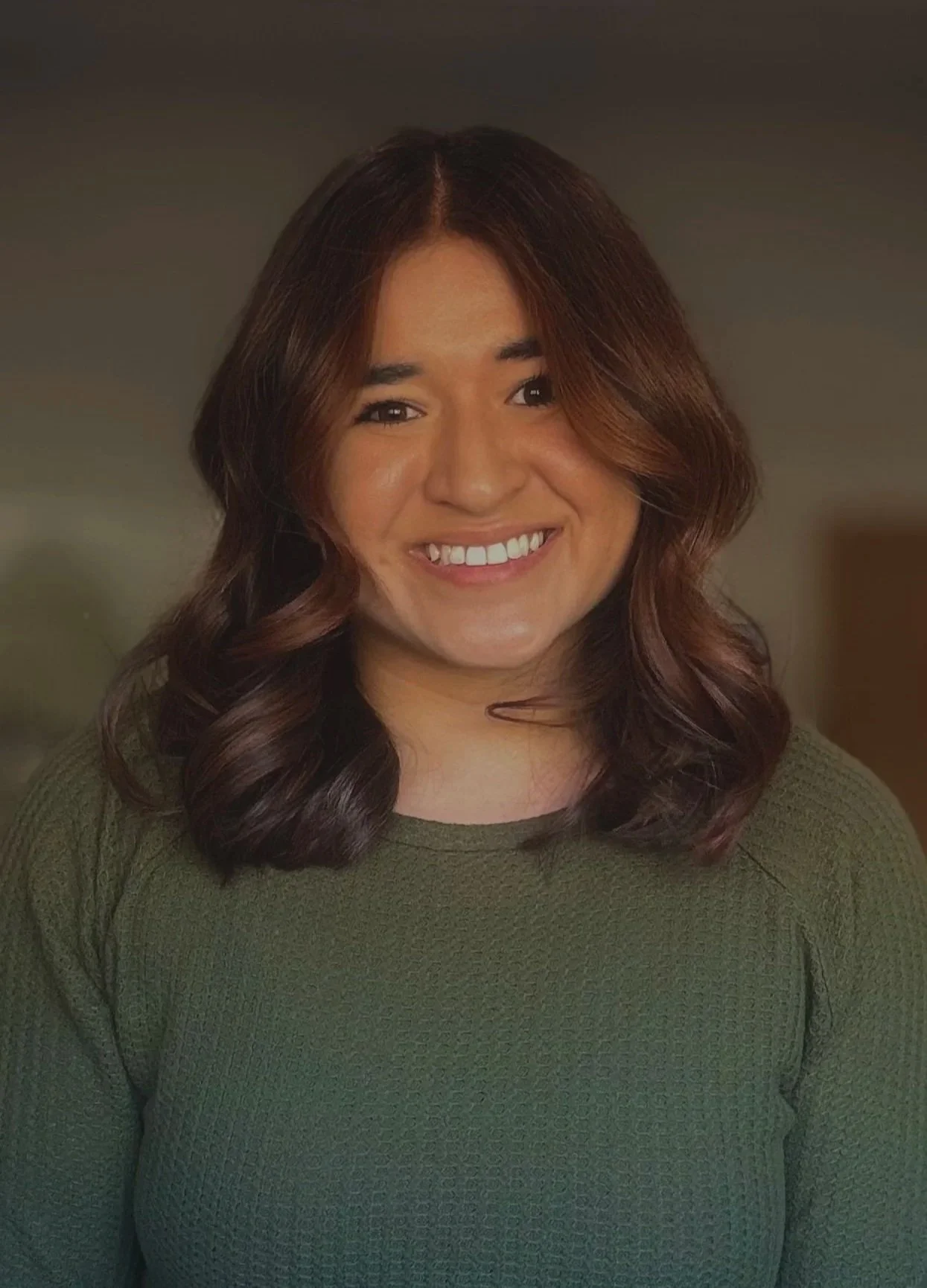 A woman with shoulder-length wavy brown hair smiling, wearing a green sweater, indoors with neutral background.