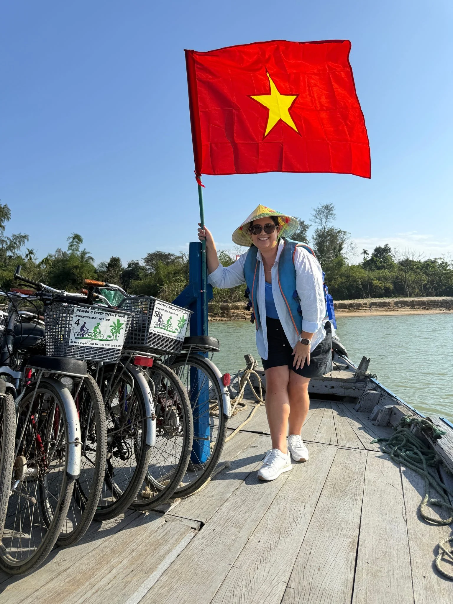 A woman standing on a wooden dock by the water, holding a red flag with a yellow star, next to a row of bicycles, wearing a traditional conical hat, sunglasses, a white shirt, black shorts, and white sneakers, smiling.