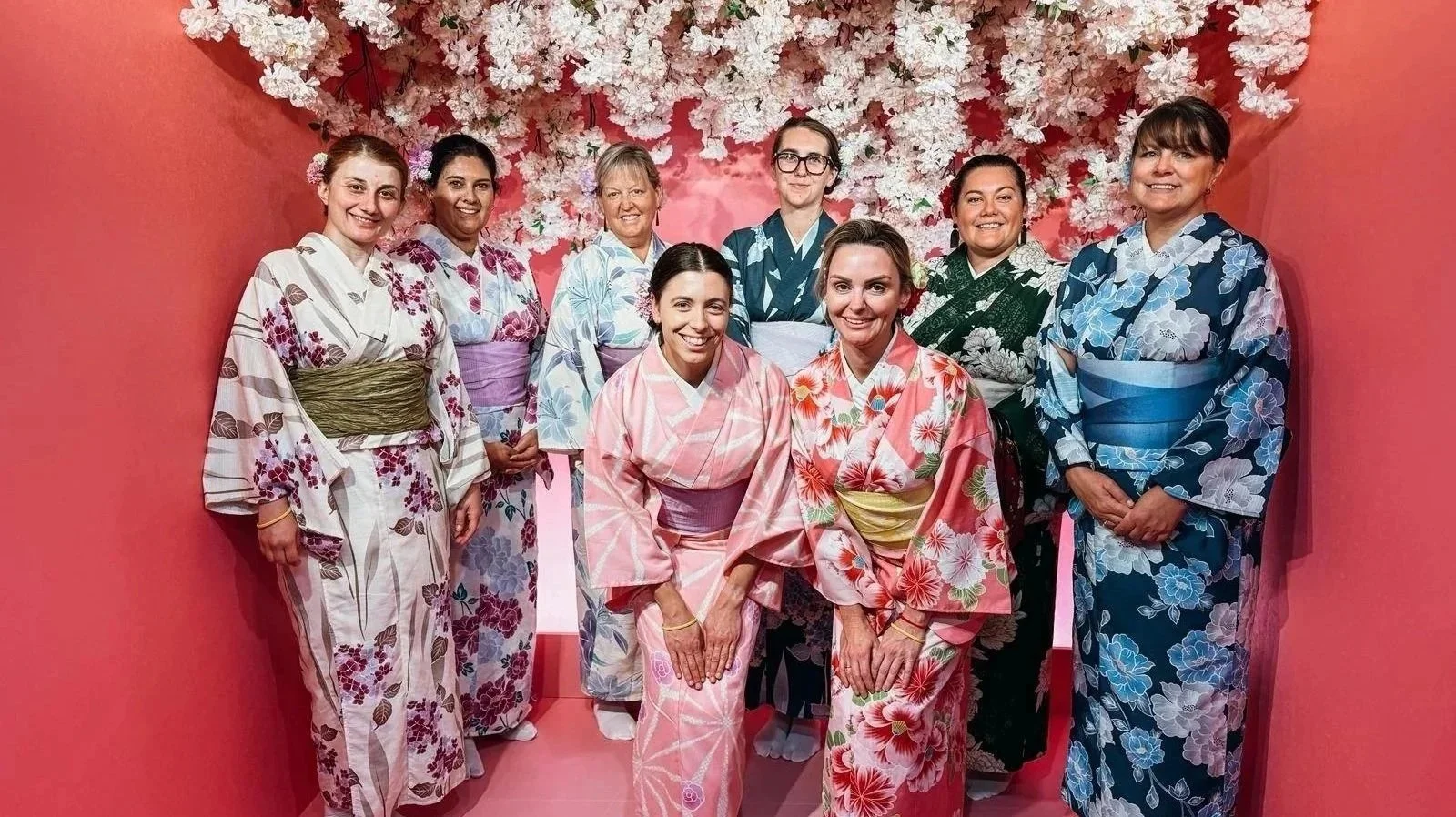 Group of women dressed in colorful kimonos, standing in a pink-walled room with a large white floral decoration on the wall behind them.