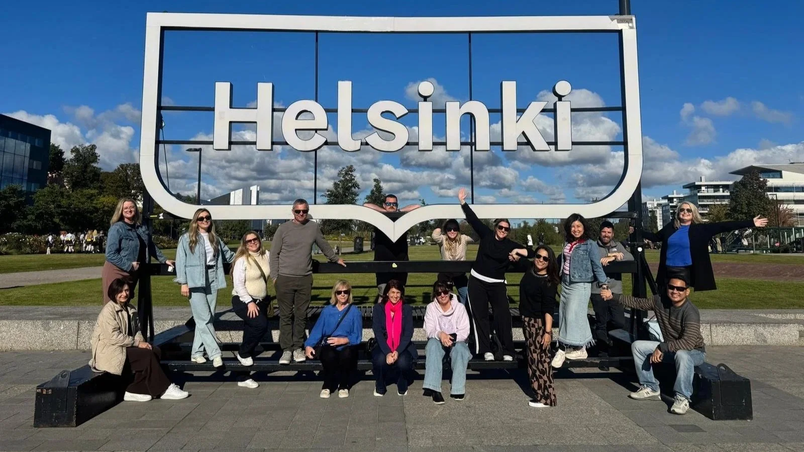 Group of people posing in front of a large Helsinki sign outdoors on a sunny day.