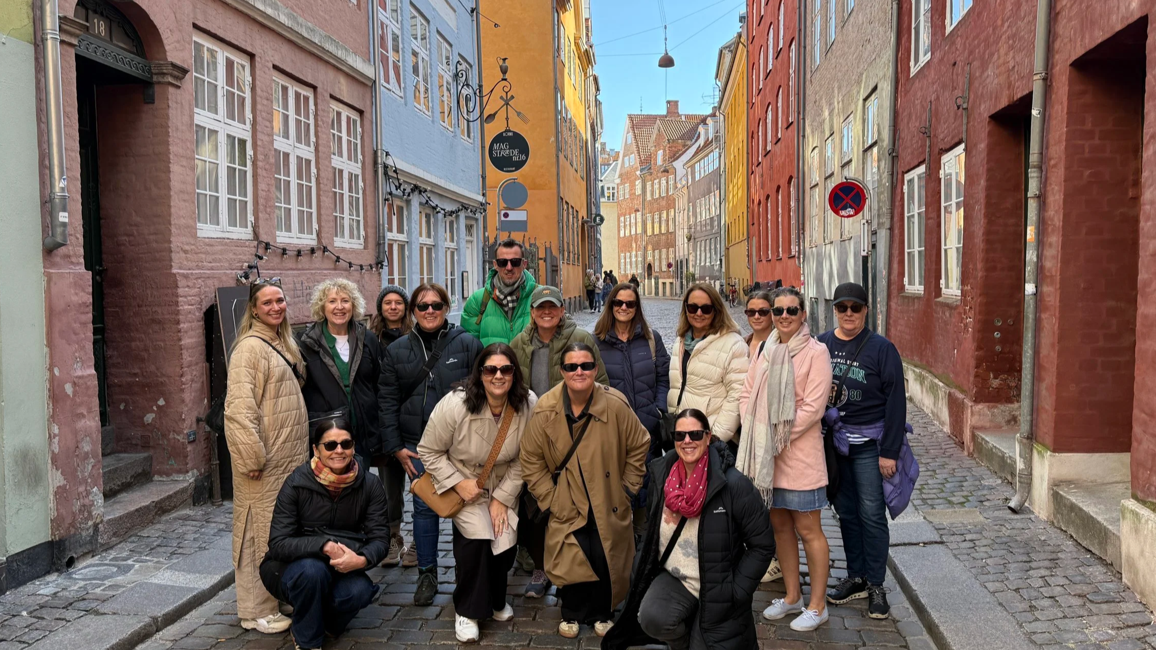 A group of tourists posing on a cobblestone street in front of colorful European buildings. The group includes women and men wearing jackets and sunglasses.