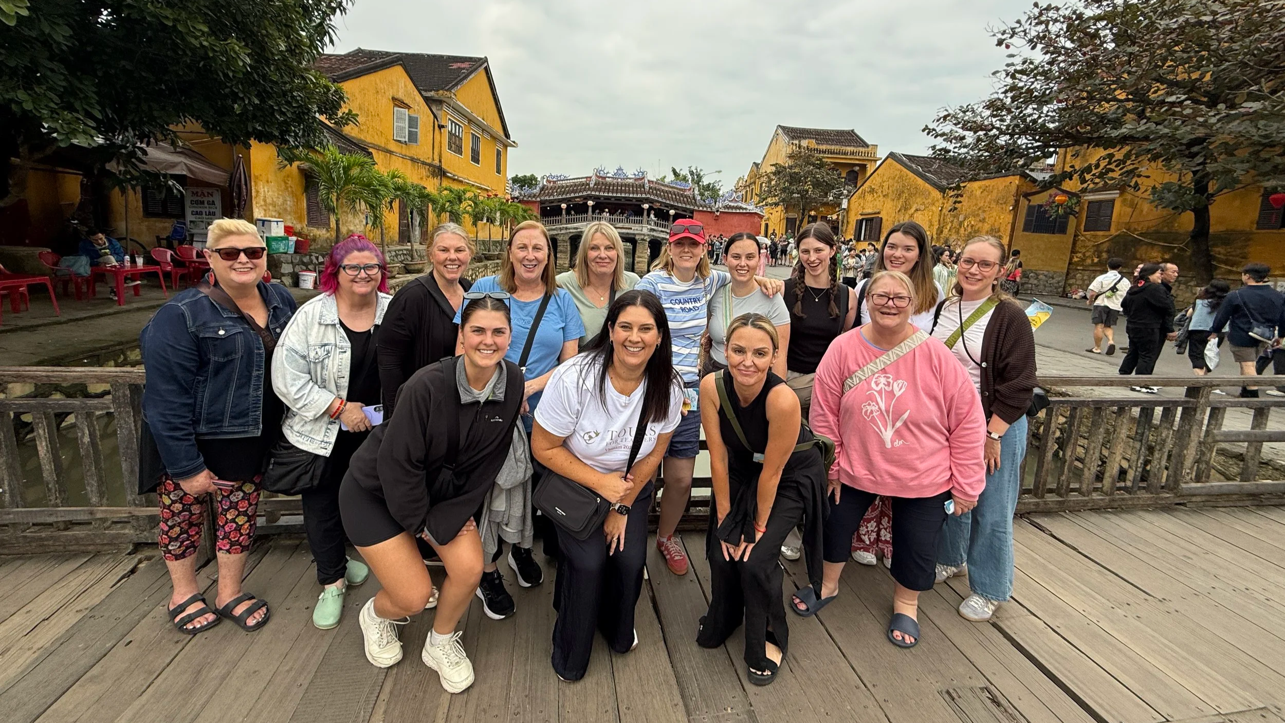 A group of 14 women posing on a wooden bridge in a tourist area with yellow buildings and a small bridge in the background.
