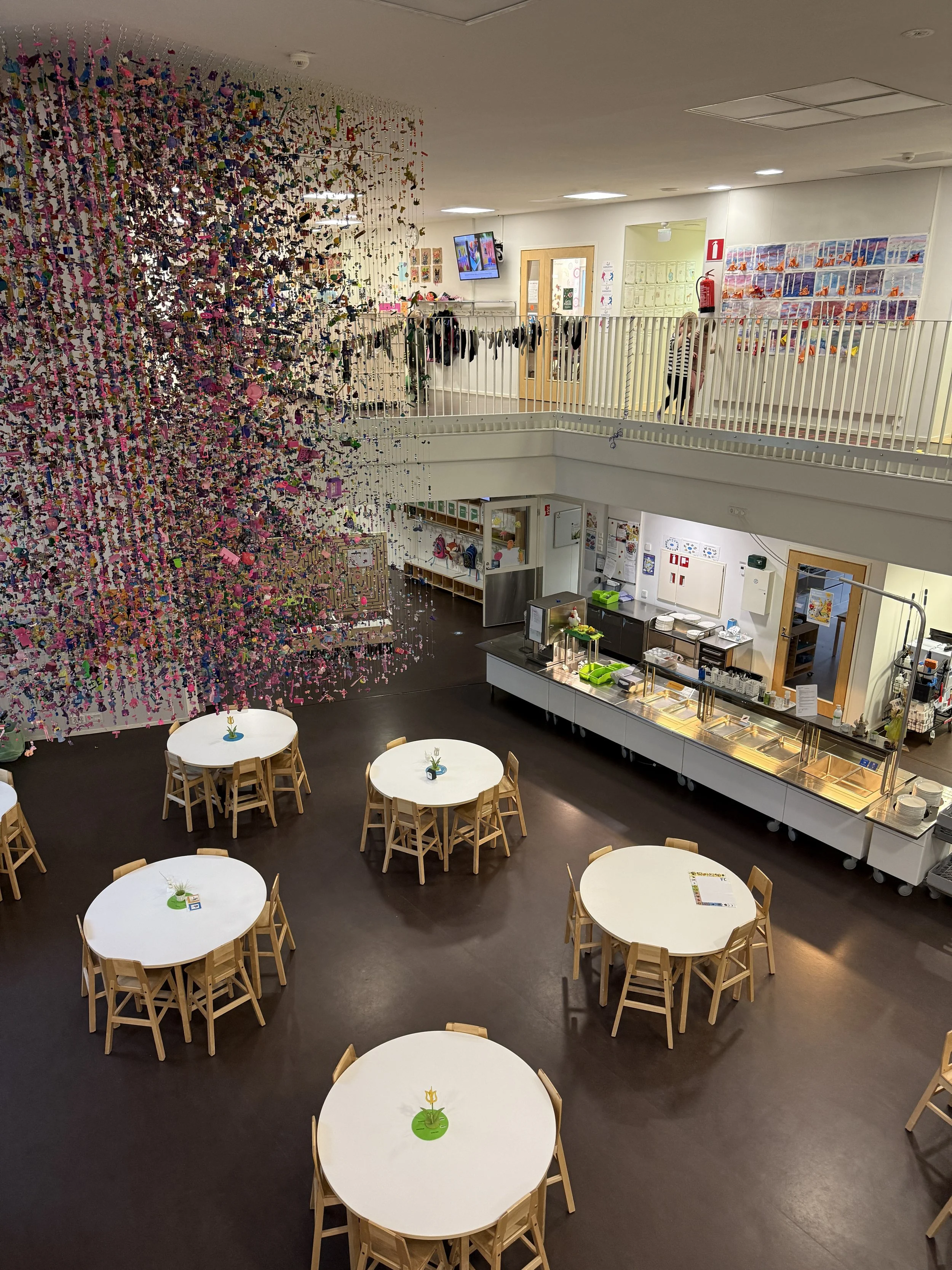 Community room with ascending levels, a colorful art installation hanging from the ceiling on the left, and round tables with wooden chairs in the foreground. The room features a cafeteria-style serving counter, lockers, and various wall decorations.