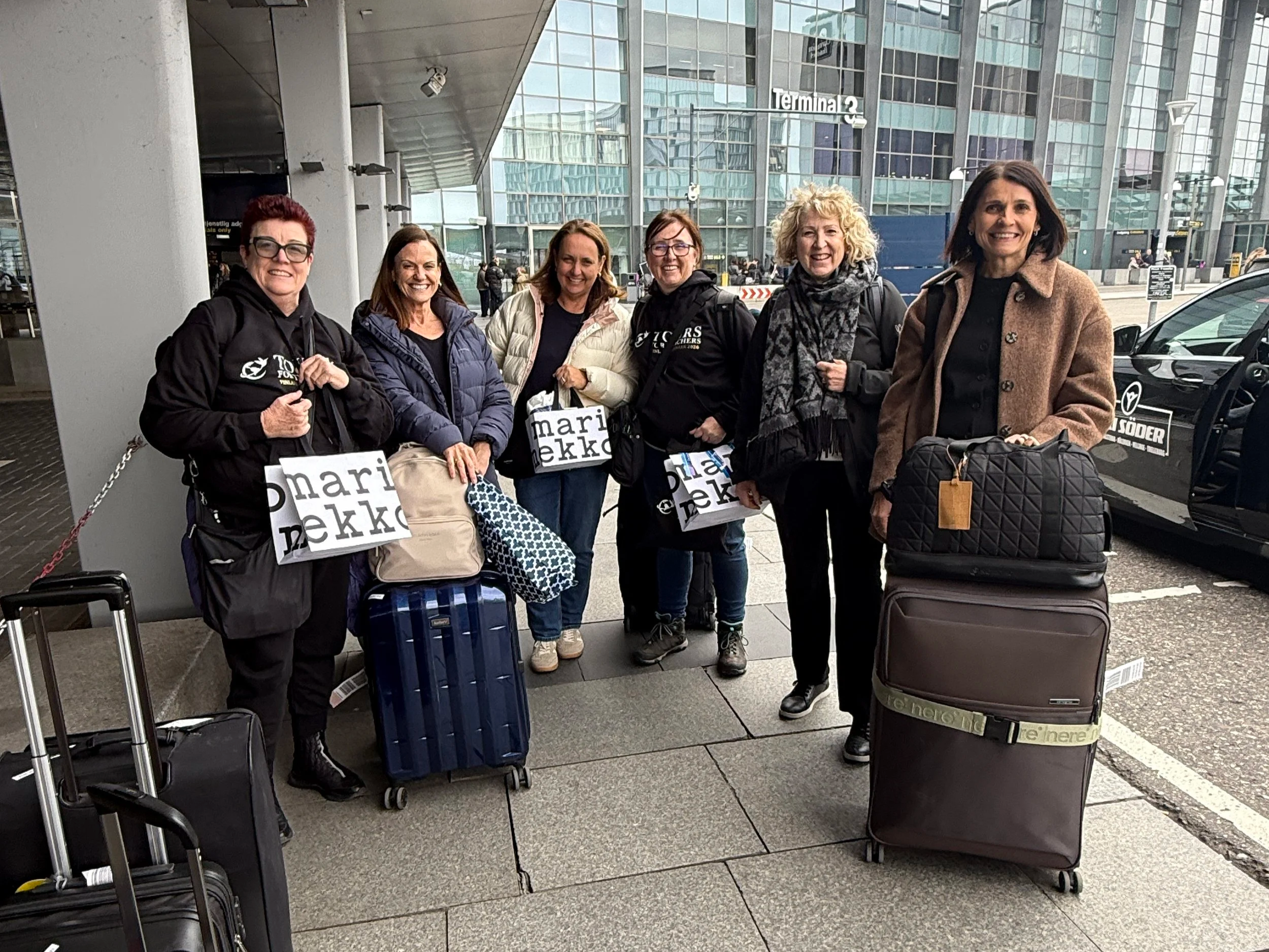 A group of six women standing outside an airport terminal with luggage, smiling at the camera.