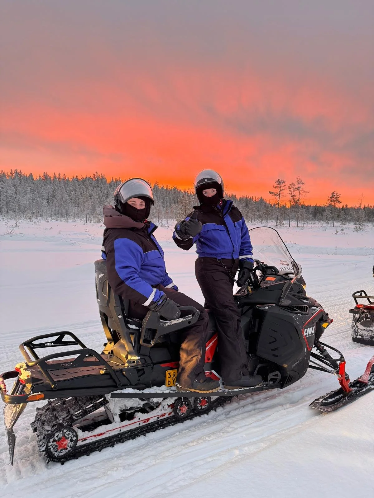 Two people in winter gear, wearing helmets and face masks, riding a snowmobile in a snowy landscape at sunset with a colorful sky and forest in the background.