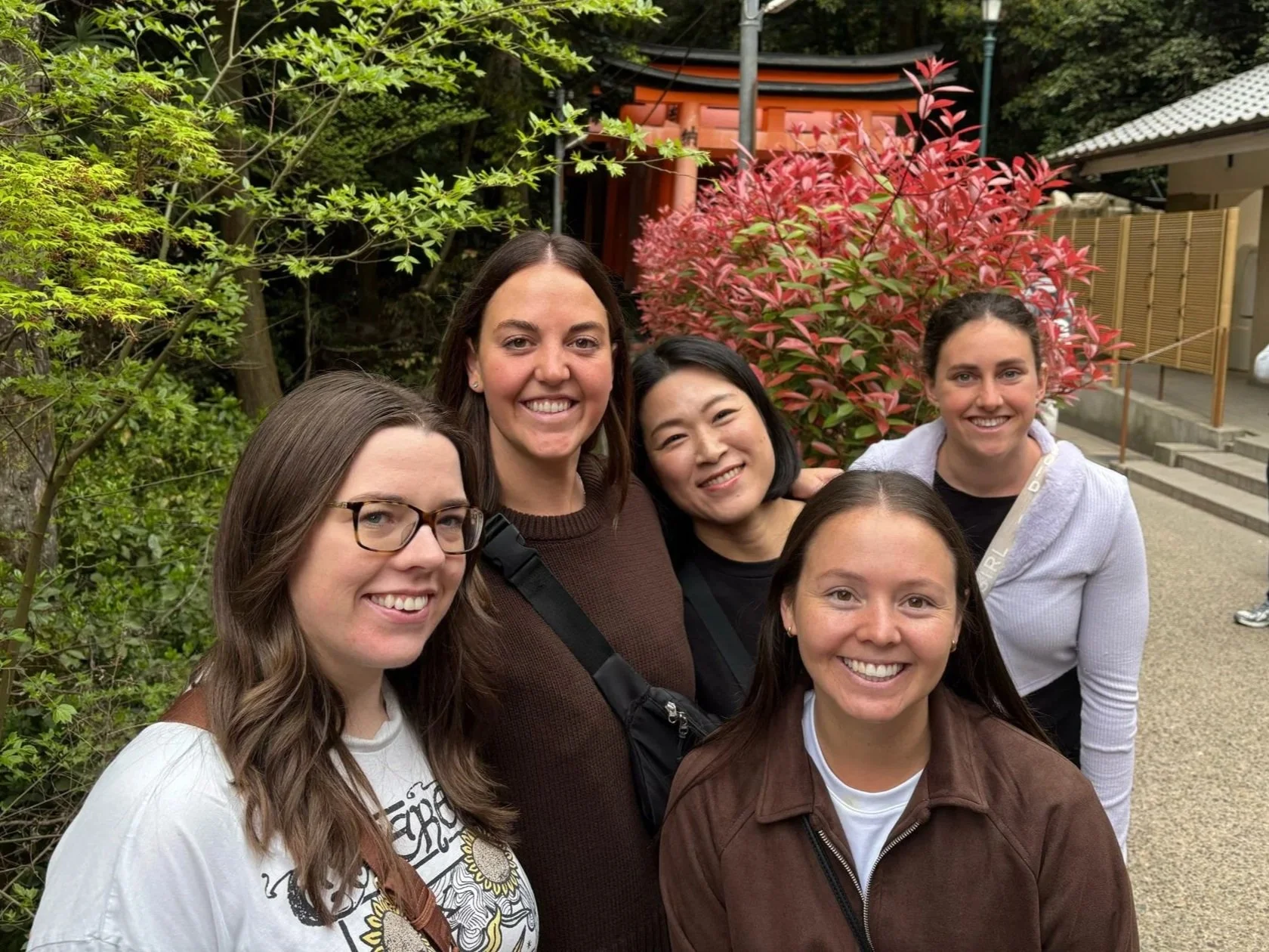 Group of five women smiling outdoors, standing in front of lush green trees and a red shrine, with steps on the side.