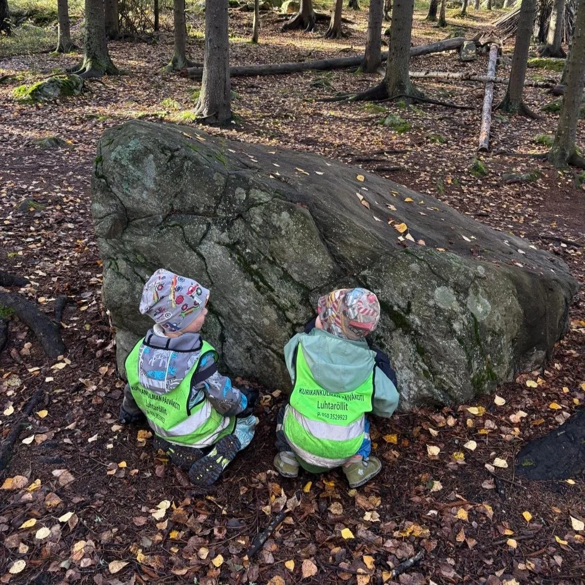 Two young children wearing bright yellow safety vests and colorful hats are crouching near a large mossy boulder in a forest, surrounded by fallen leaves and trees.