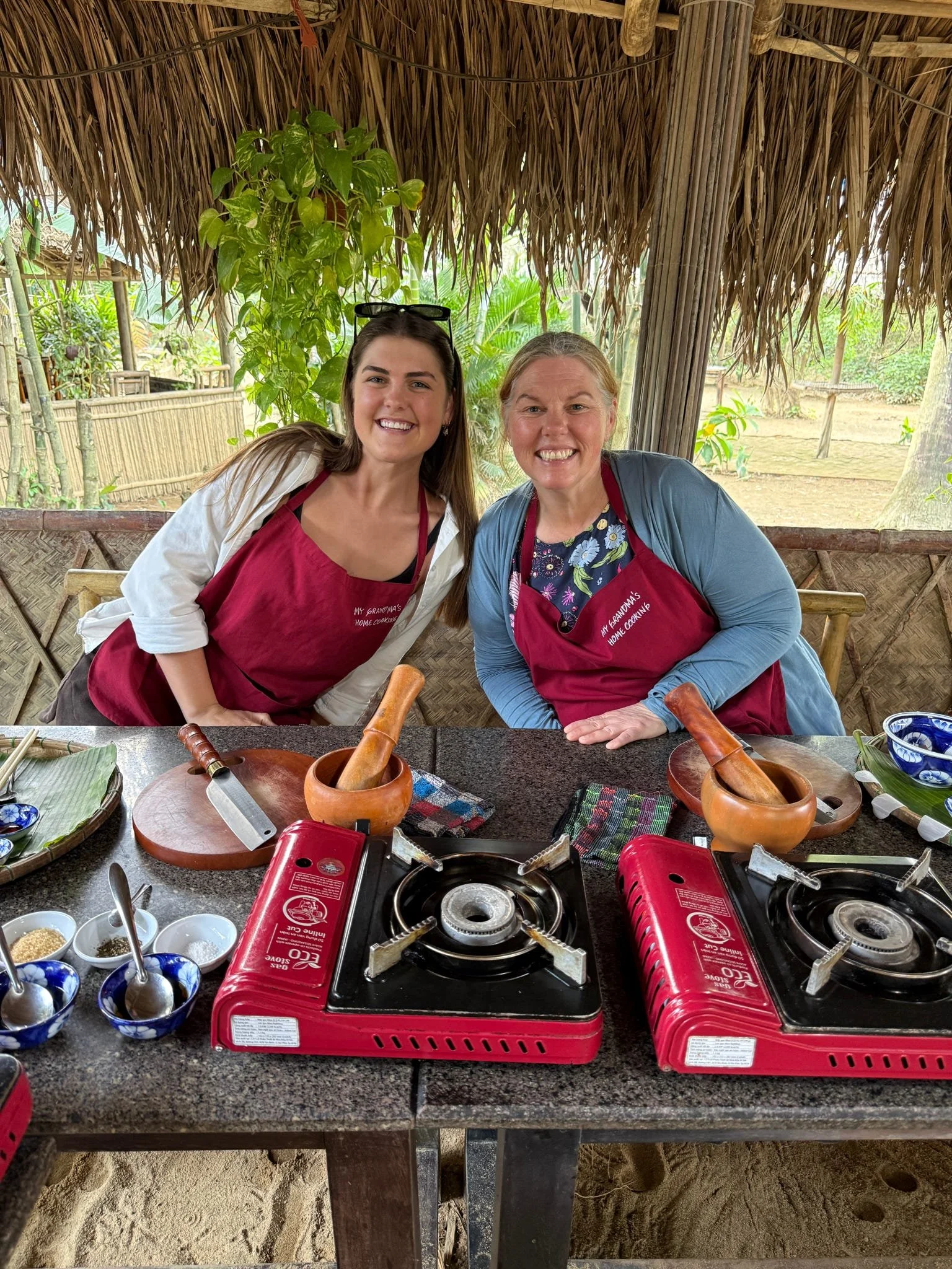 Two women in aprons smiling at a cooking setup with two portable gas stoves, bowls, utensils, and ingredients on a granite counter under a thatched roof, surrounded by tropical greenery.