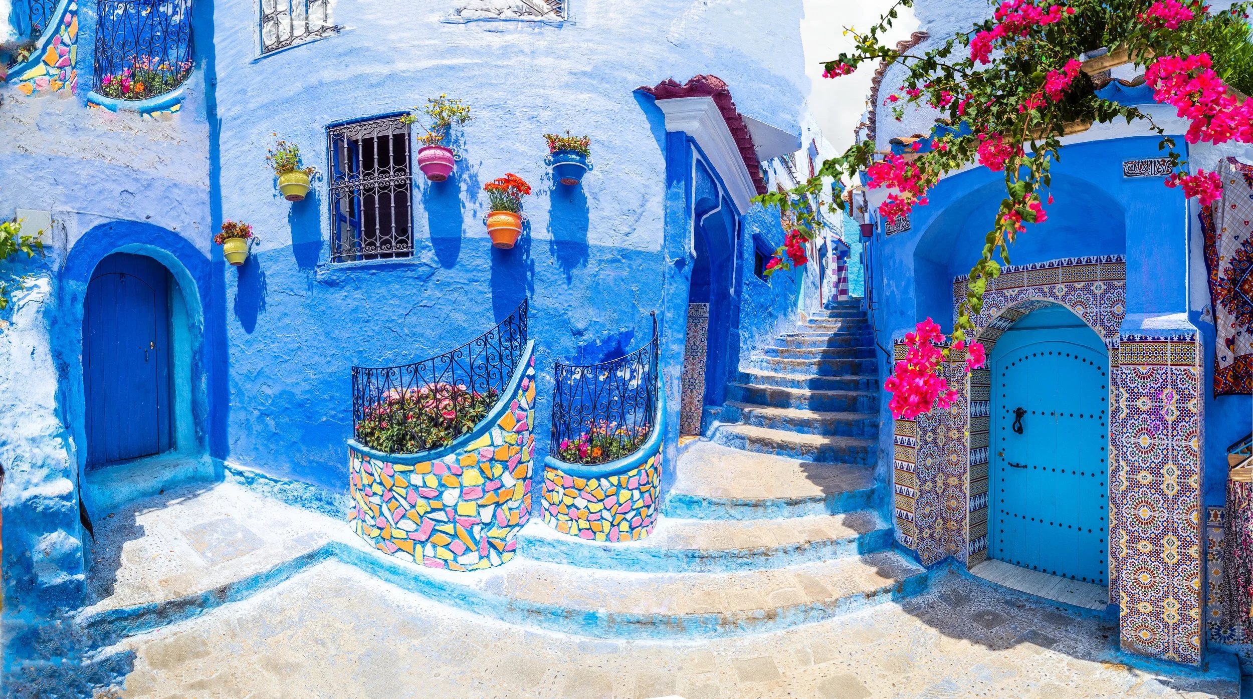 Colorful blue-painted building with decorative tiles, potted plants, flowering vines, and steps leading up to a courtyard, capturing a vibrant Mediterranean or North African street scene.