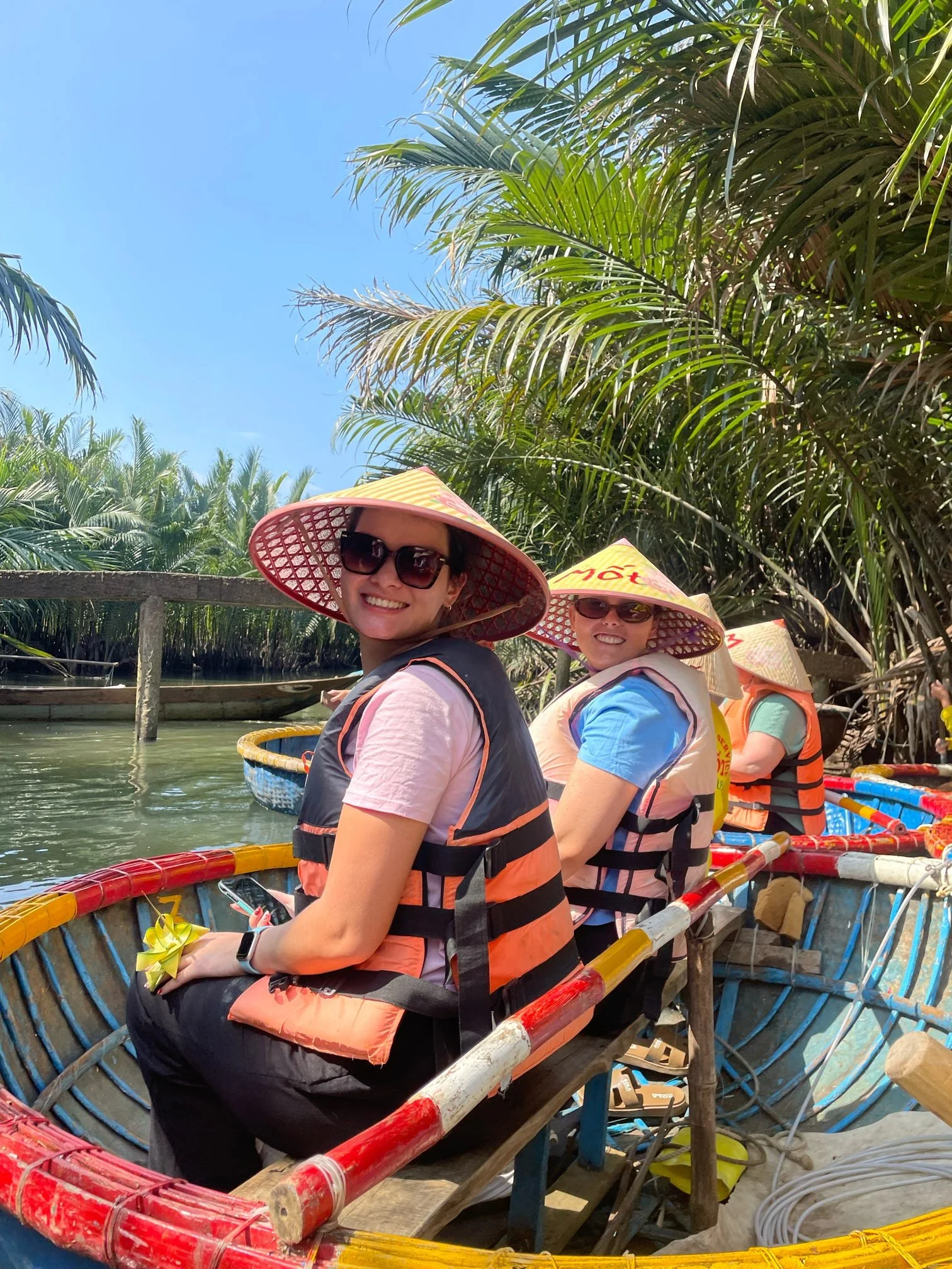 Two smiling women wearing sun hats and sunglasses sitting on a boat made of bamboo, surrounded by lush greenery and palm trees, on a sunny day.