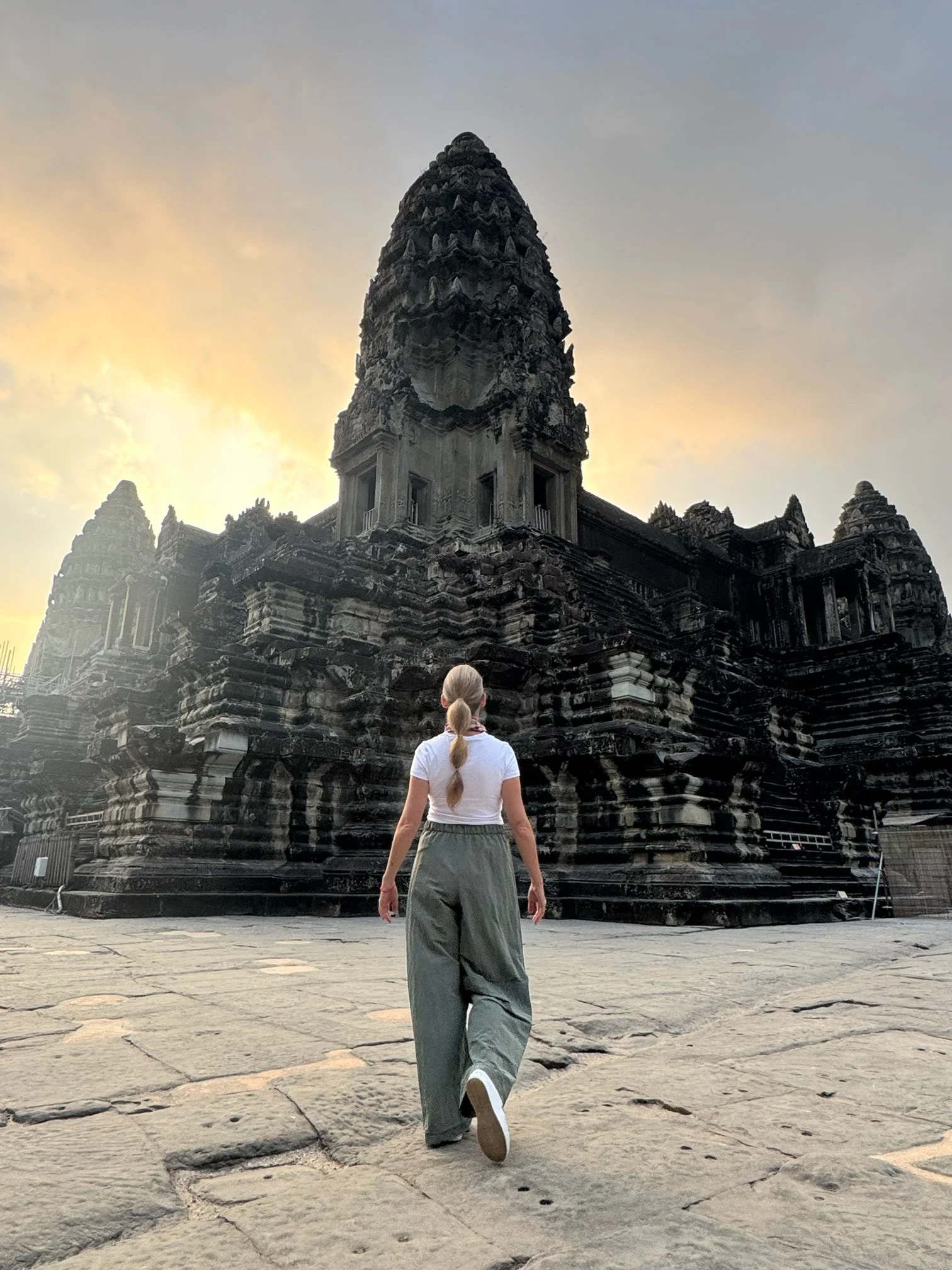 A woman with blonde hair tied in a ponytail, wearing a white t-shirt and loose green pants, walks toward an ancient temple with intricate stone carvings at sunset.