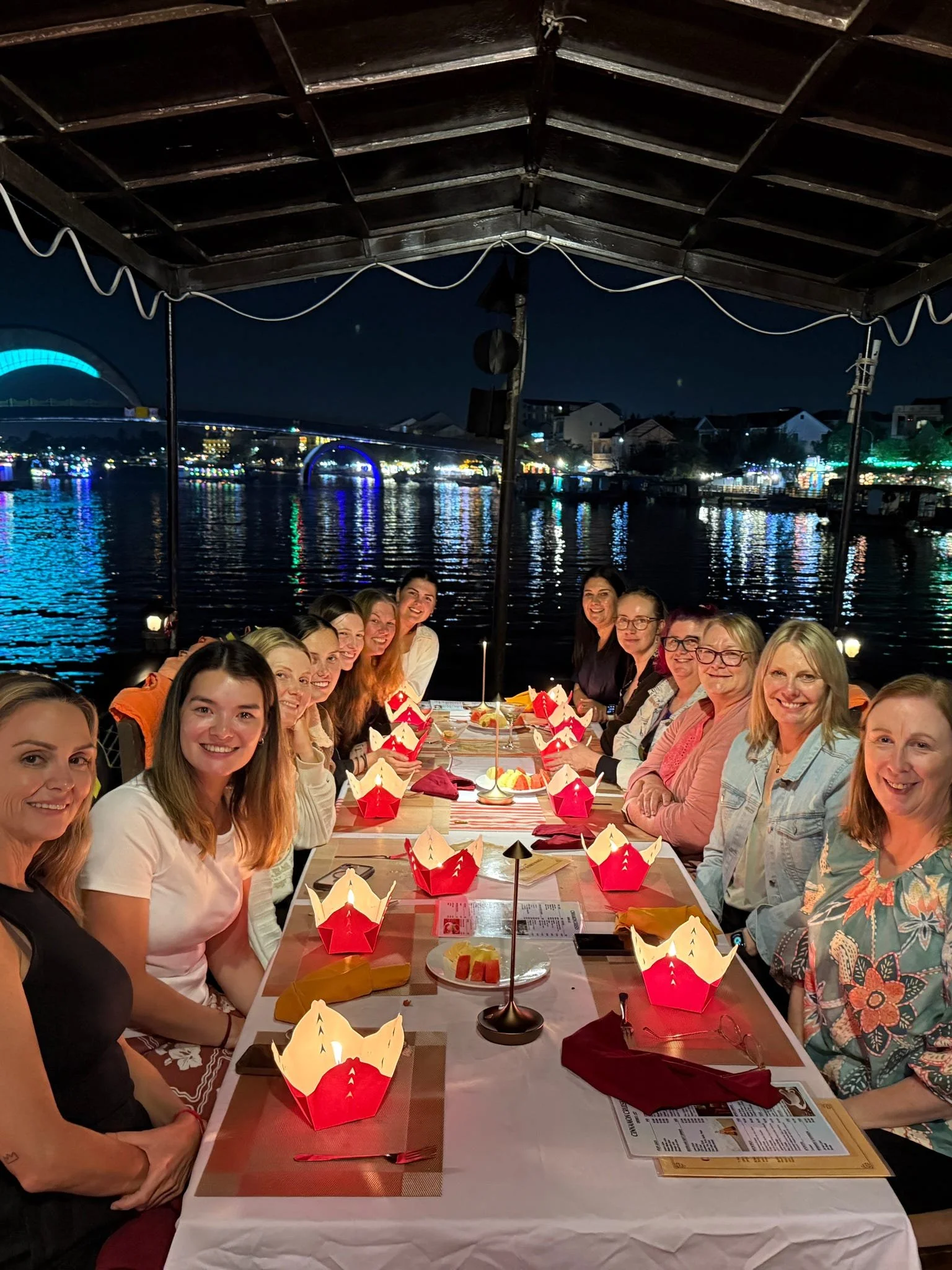People gathered around a dinner table at night with lights reflecting on the water behind them, enjoying a meal together outdoors under a canopy.