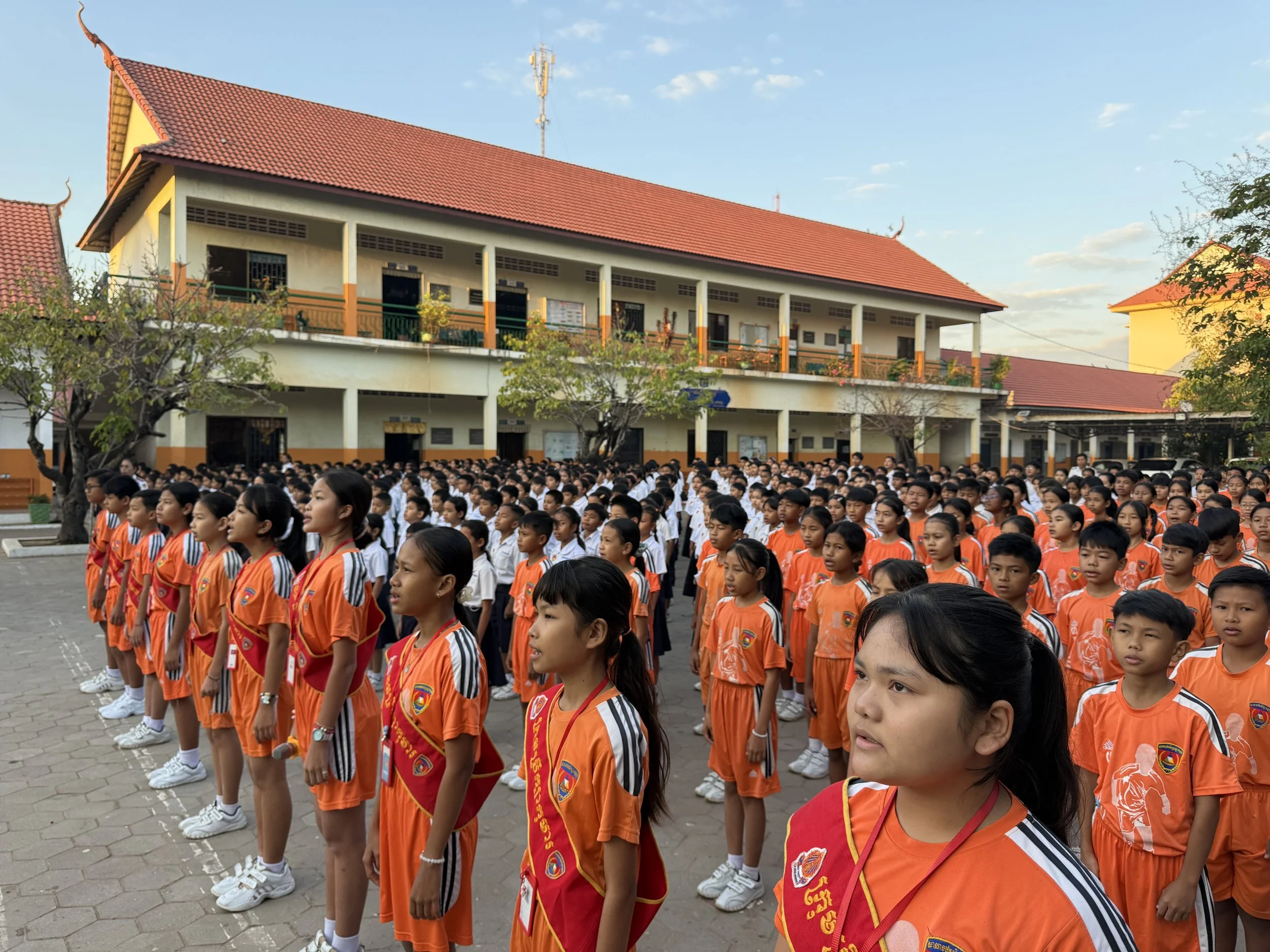 School children standing in orderly rows outside school building, dressed in orange sports uniforms with badges, some wearing red sashes, under trees in bright daylight.