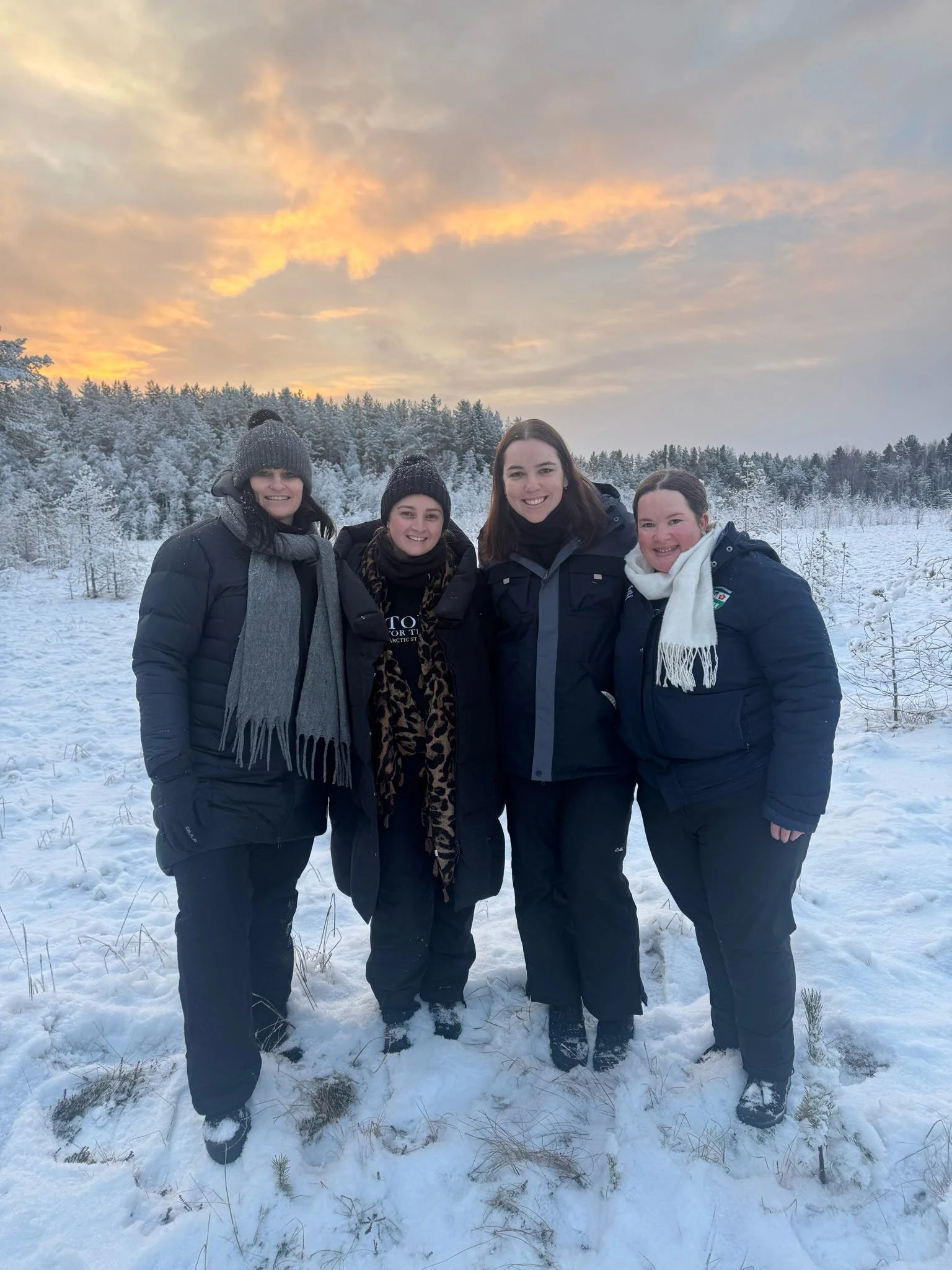 Four women standing together in a snowy field with a forest and a sunset sky in the background, all dressed in winter clothing.