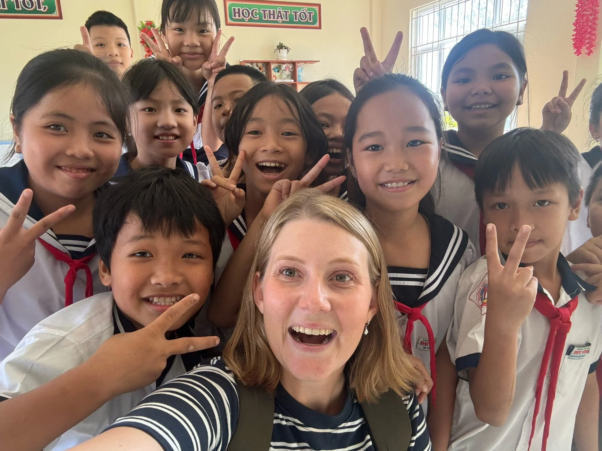 A group of children and a woman taking a selfie together inside a classroom. The children are smiling and making peace signs, and there are educational posters on the wall.