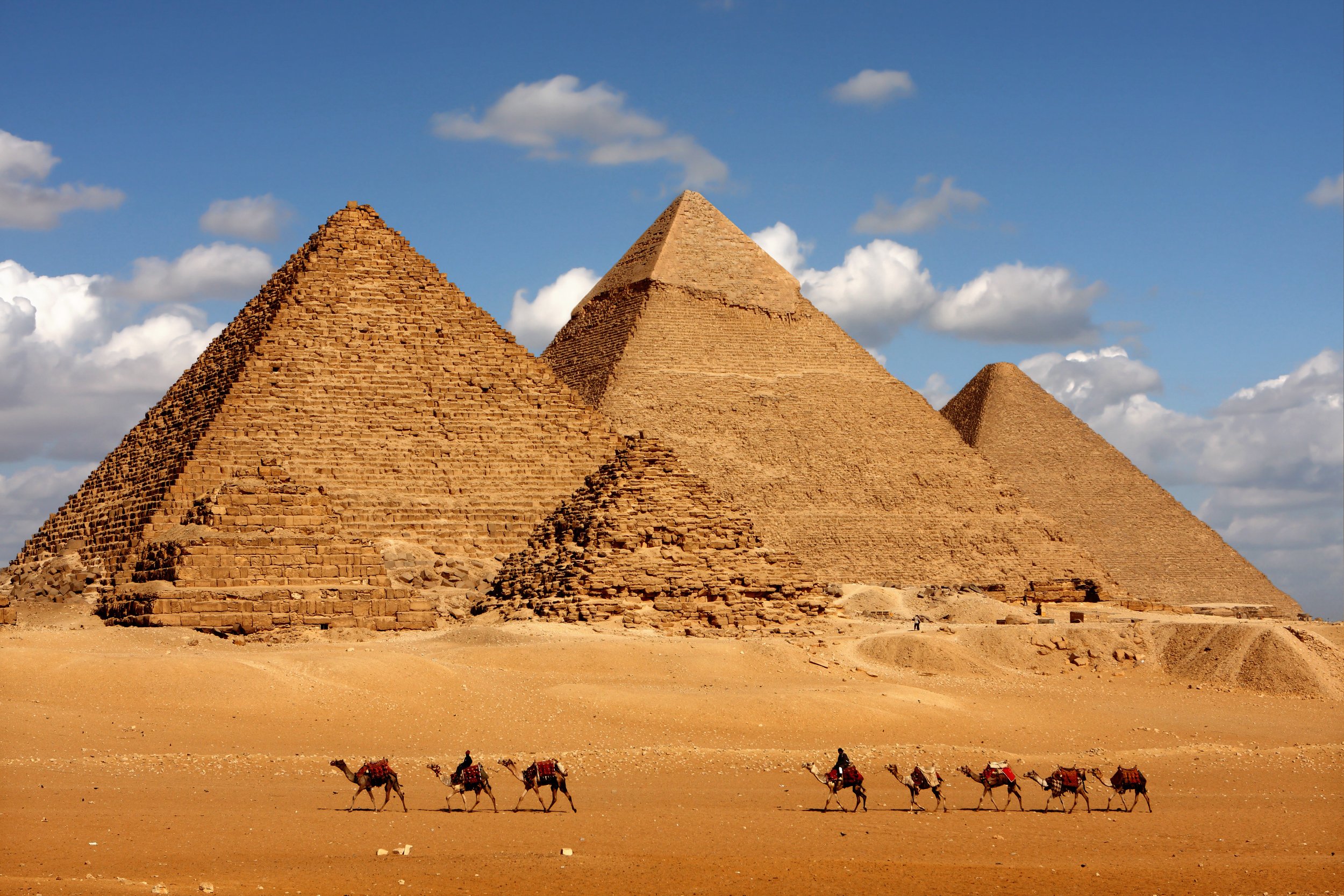 A desert scene with three large pyramids, a camel caravan in front, and a cloudy blue sky above.